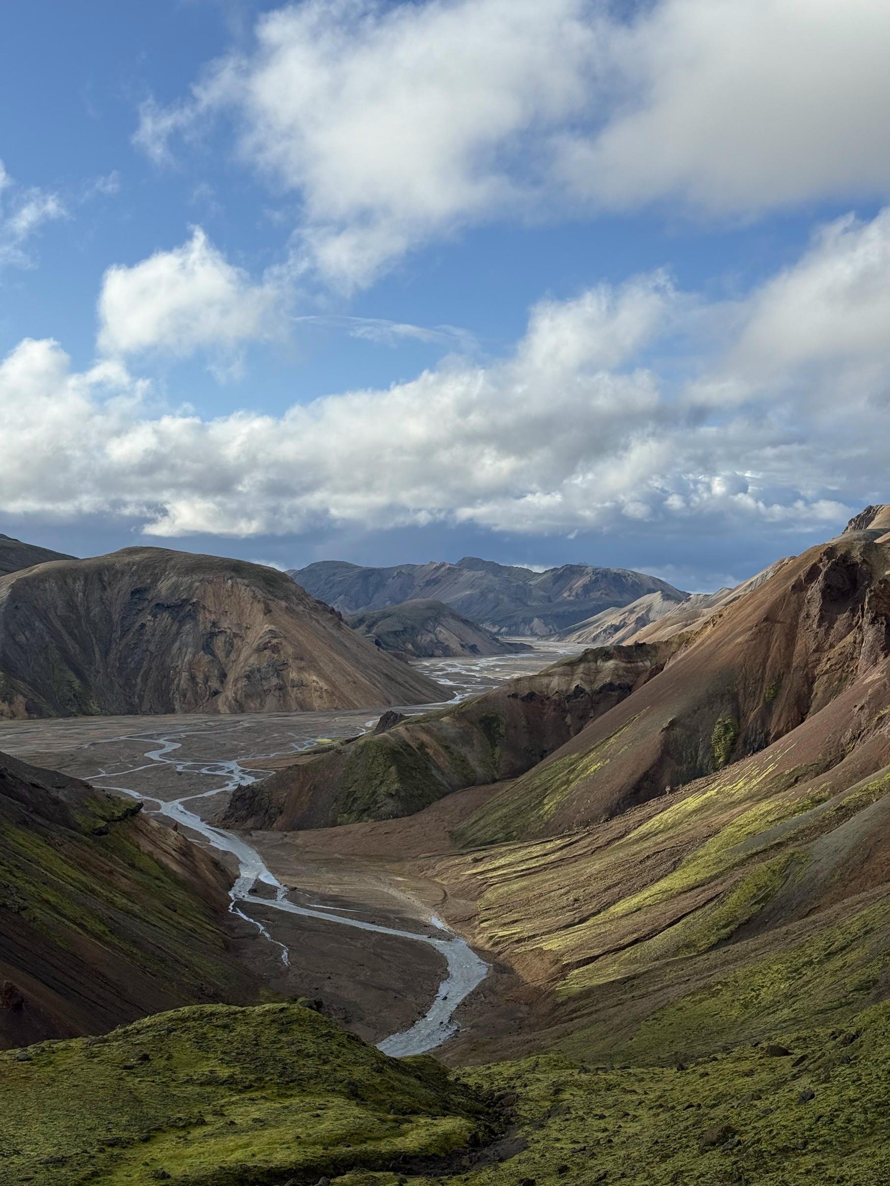 Ein malerischer Blick auf einen gewundenen Fluss, der sich durch grüne, moosbewachsene Hügel und felsige Berge schlängelt, unter einem teilweise bewölkten blauen Himmel.