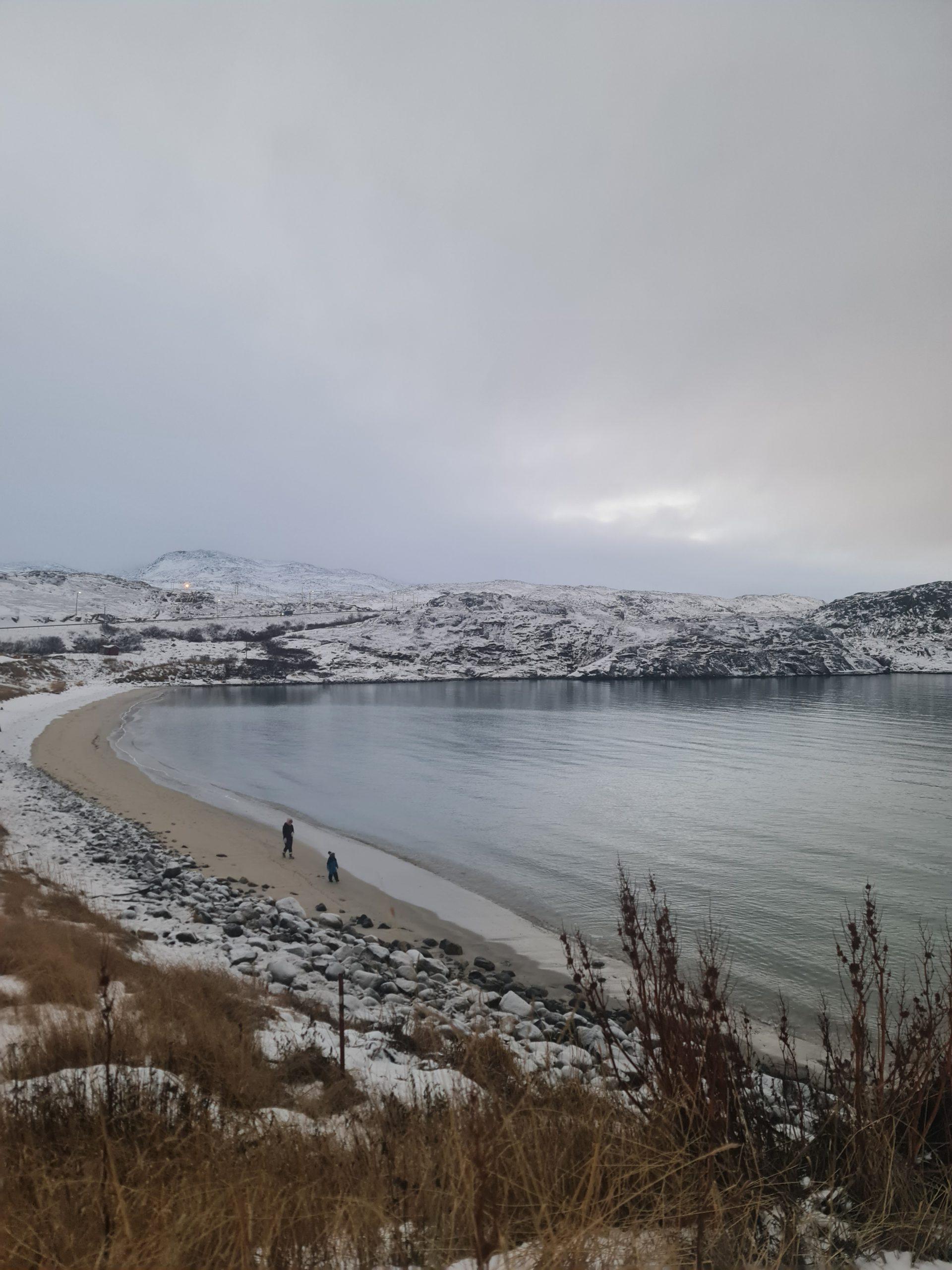 Eine ruhige, verschneite Strandszene mit zwei Personen, die am Ufer spazieren gehen. Schneebedeckte Hügel umgeben die Bucht, und trockenes Gras säumt den Vordergrund unter einem bewölkten Himmel.