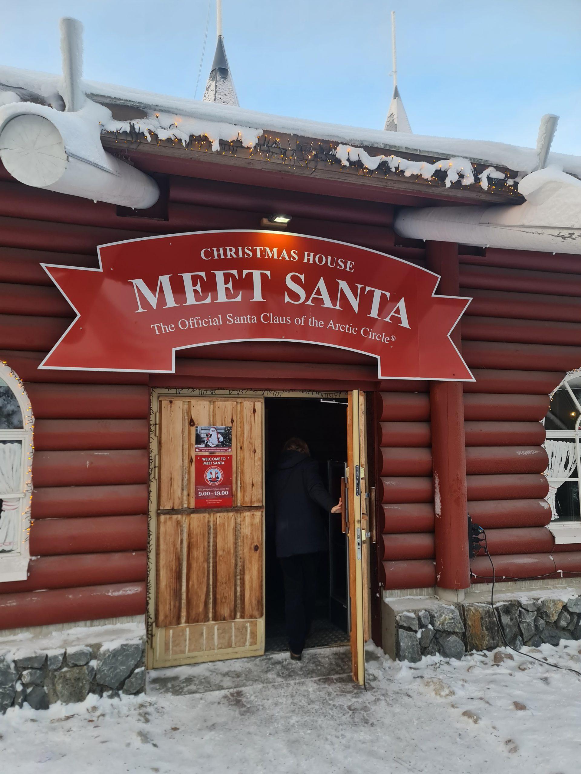 Eine Person betritt eine Blockhütte mit einem roten Schild über der Tür, auf dem "Christmas House Meet Santa - The Official Santa Claus of the Arctic Circle" steht. Auf dem Dach und dem Boden liegt Schnee.