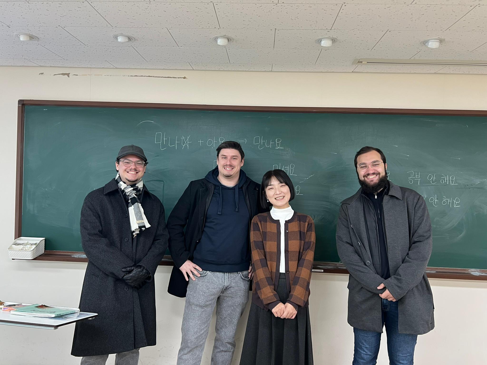 Vier Personen, drei Männer und eine Frau, stehen lächelnd vor einer Kreidetafel mit koreanischer Schrift in einem Klassenzimmer. Sie sind warm gekleidet, was auf eine kalte Jahreszeit hindeutet, und wirken entspannt und freundlich.