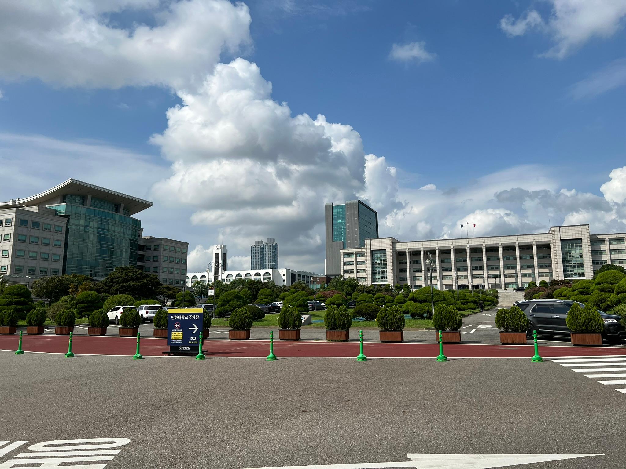 Ein großer, moderner Universitätscampus mit zwei markanten Gebäuden, gepflegten Büschen, geparkten Autos und einem strahlend blauen Himmel mit Schäfchenwolken über dem Kopf. Ein Schild und grüne Schranken markieren den Eingangsbereich.