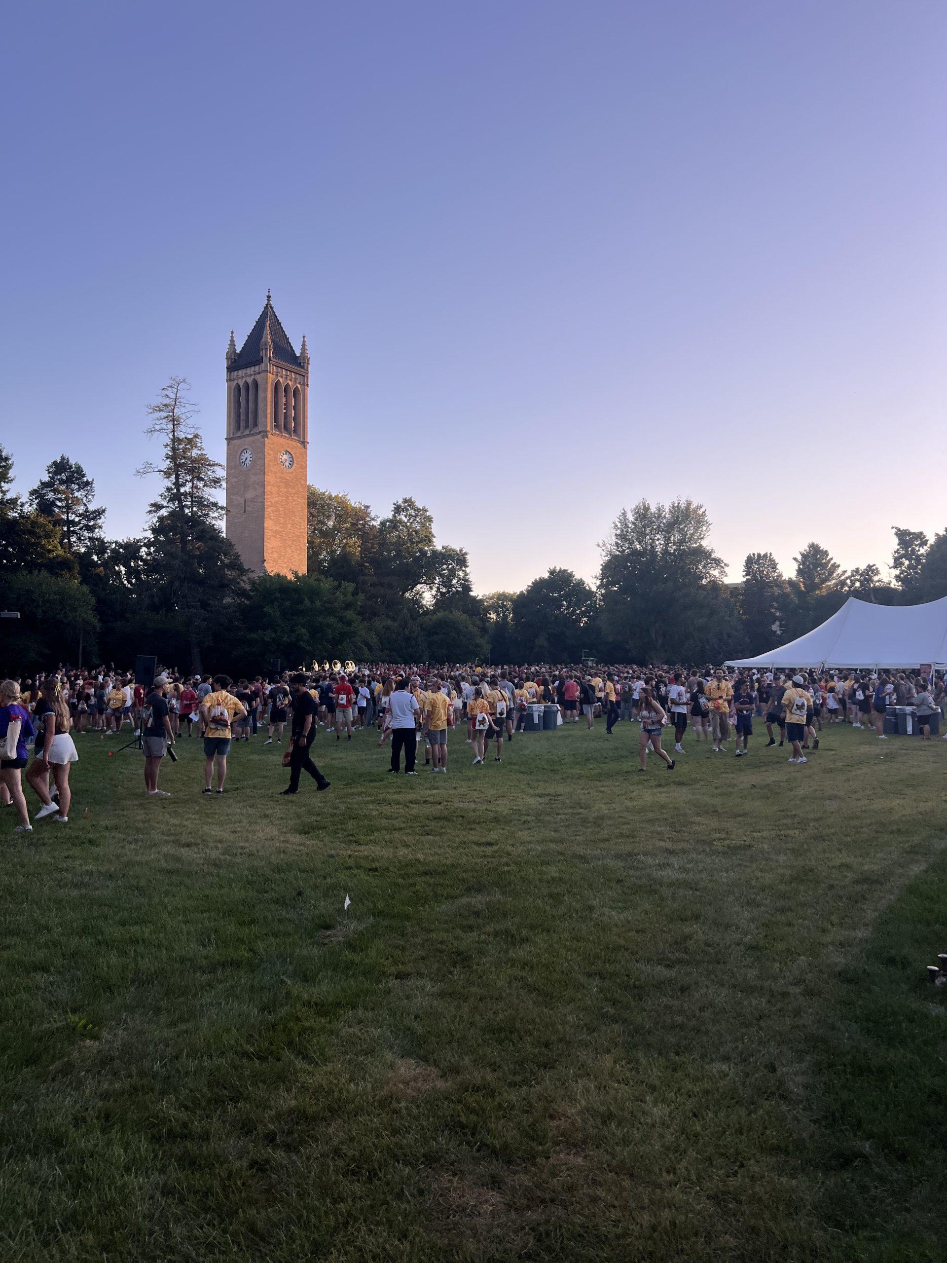 Eine Menschenmenge versammelt sich bei Sonnenuntergang auf einer Wiese in der Nähe eines hohen Uhrenturms, mit Bäumen im Hintergrund und einem großen weißen Zelt auf der rechten Seite. Der Himmel ist klar, und die Menschen tragen legere Sommerkleidung.