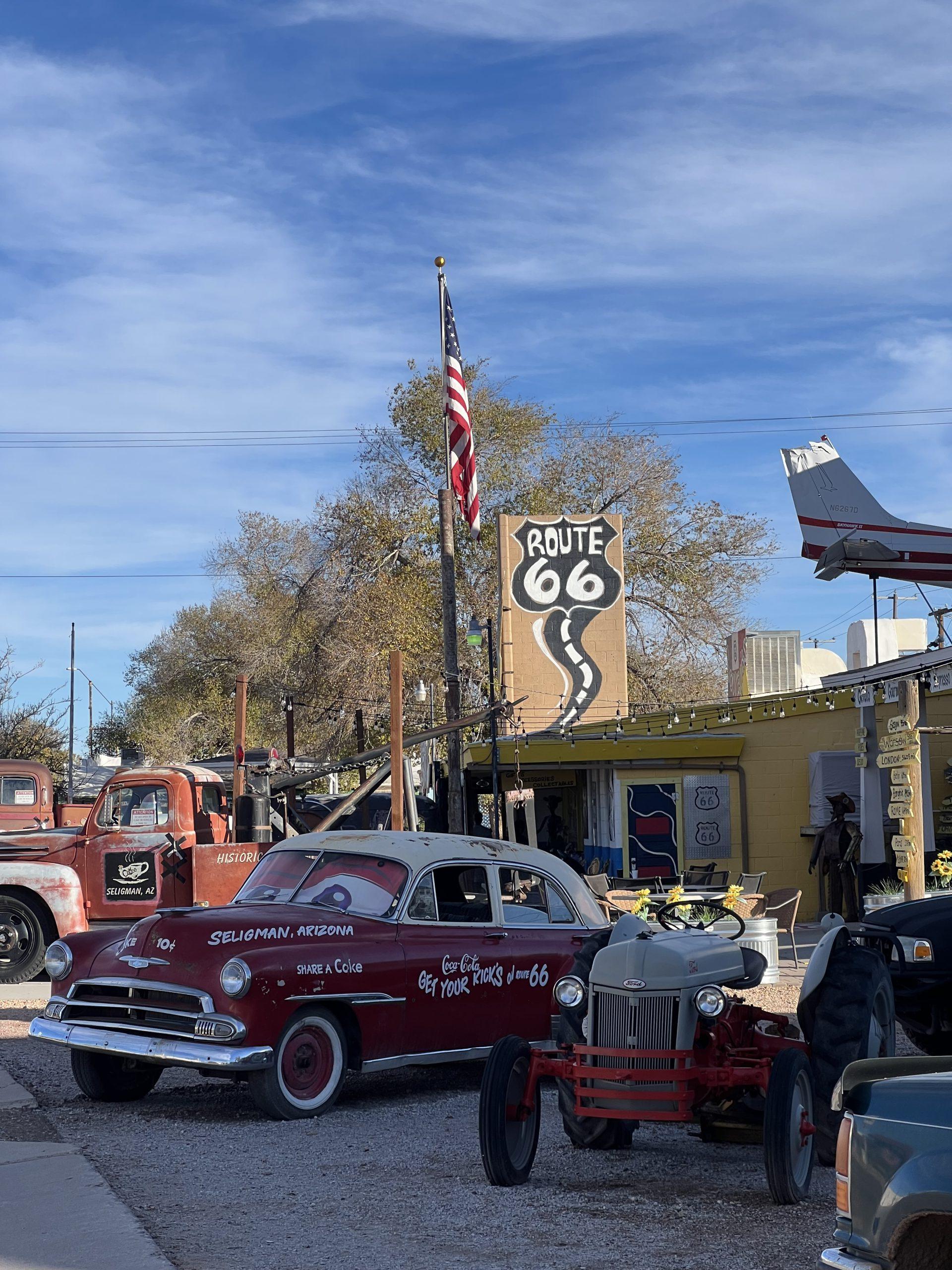Ein roter Oldtimer und ein alter Traktor stehen vor einem Retro-Café im Stil der Route 66 in Seligman, Arizona. Ein großes Route-66-Schild, eine amerikanische Flagge und alte Zapfsäulen sind zu sehen, und in der Nähe stehen weitere klassische Fahrzeuge.