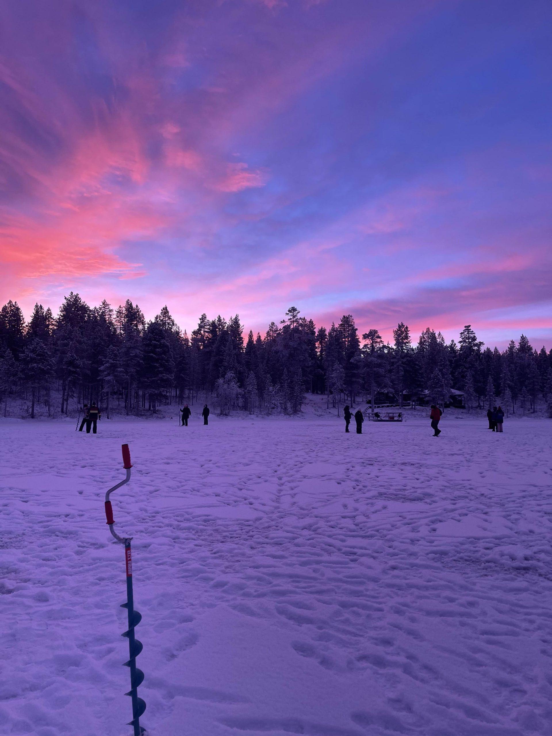 Menschen stehen in einer schneebedeckten, von Bäumen umgebenen Landschaft bei Sonnenuntergang, wobei der Himmel rosa und violett leuchtet. Im Vordergrund ist ein Eisbohrer zu sehen, der auf Eisfischen hindeutet.