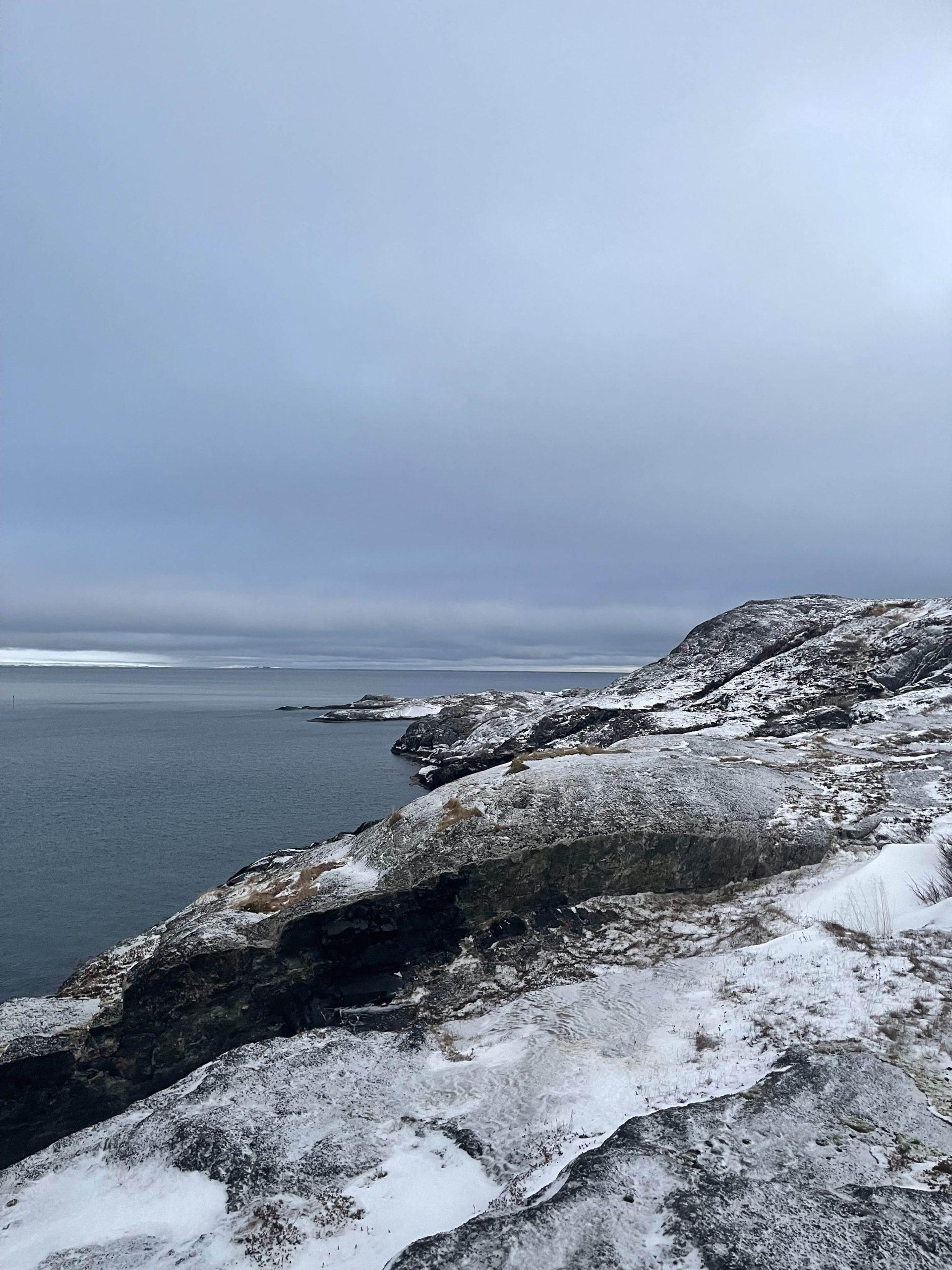 Eine felsige, schneebedeckte Küste erstreckt sich in der Ferne neben ruhigem, graublauem Wasser unter einem bedeckten Himmel. Entlang der Küste sind niedrige Hügel und spärliche Vegetation zu sehen.