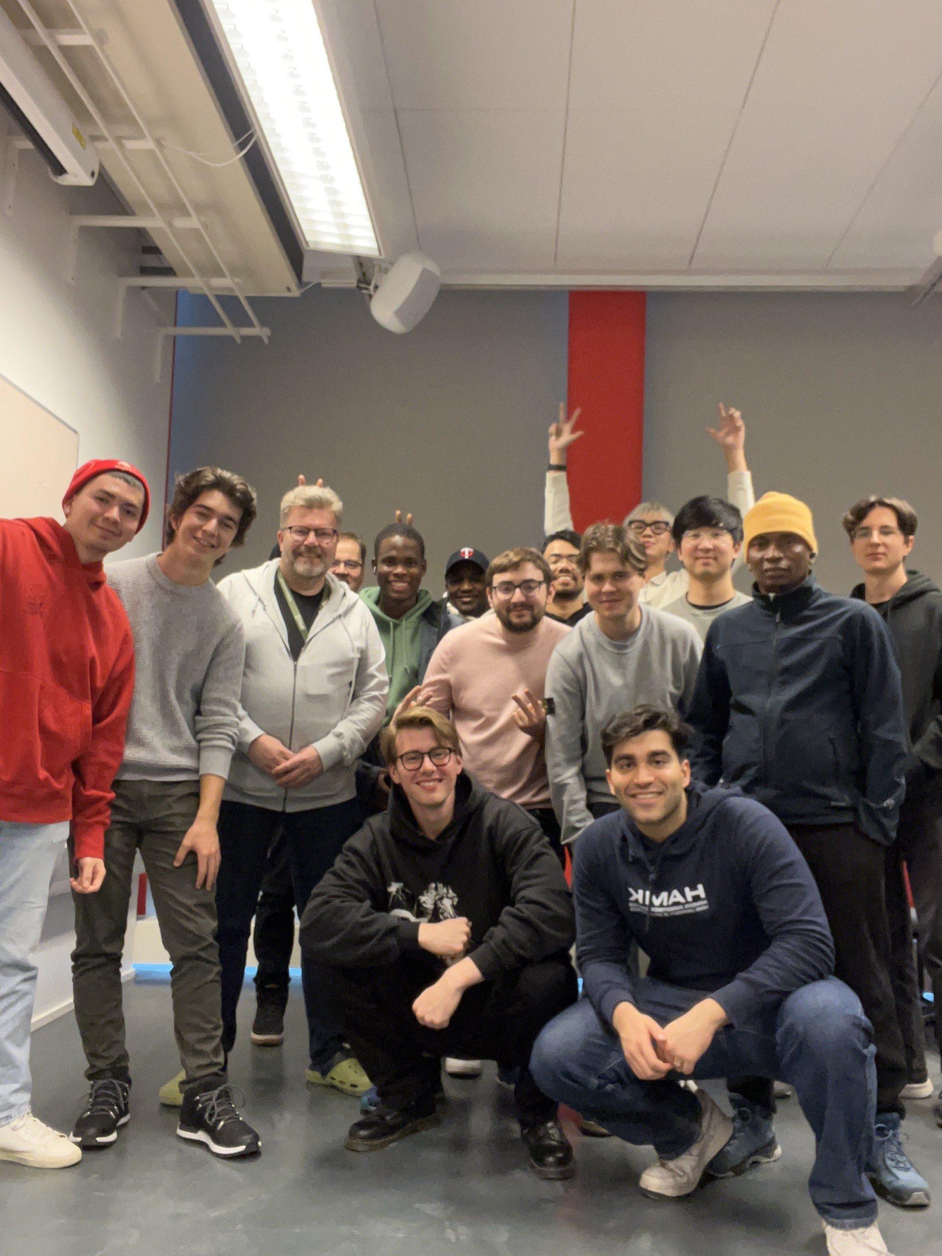 Eine Gruppe von sechzehn Personen, zumeist junge Männer, posiert und lächelt gemeinsam in einem gut beleuchteten Innenraum mit grauem Boden, weißen Wänden und einem roten vertikalen Streifen im Hintergrund. Einige machen spielerische Gesten.