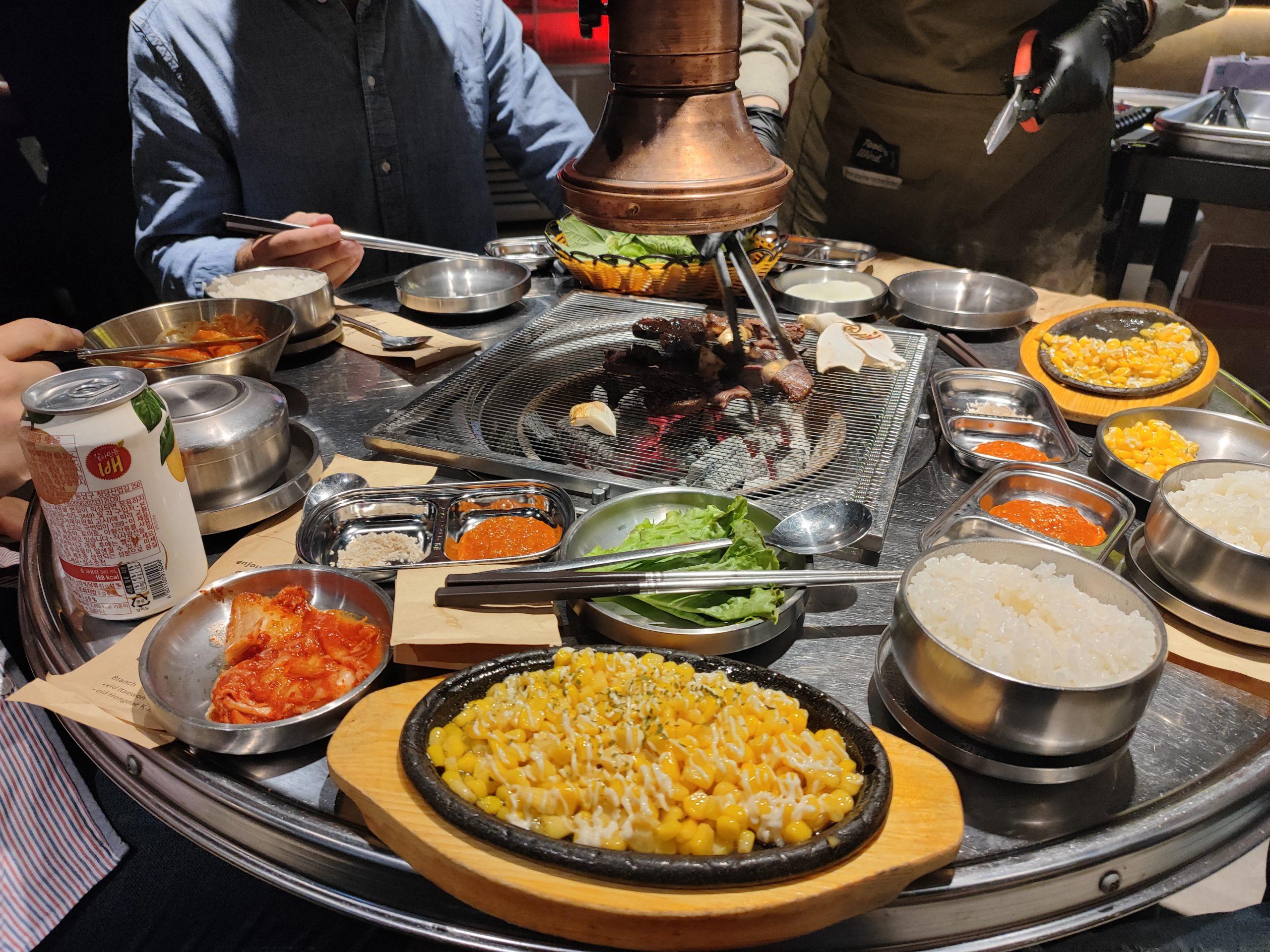 Eine Gruppe teilt sich ein koreanisches Barbecue an einem runden Tisch und grillt Fleisch und Gemüse über einem zentralen Grill. Beilagen, Reis, Maiskäse, Soßen, Kimchi und Getränke sind um den Tisch herum angeordnet. Die Hände halten Stäbchen und Utensilien.