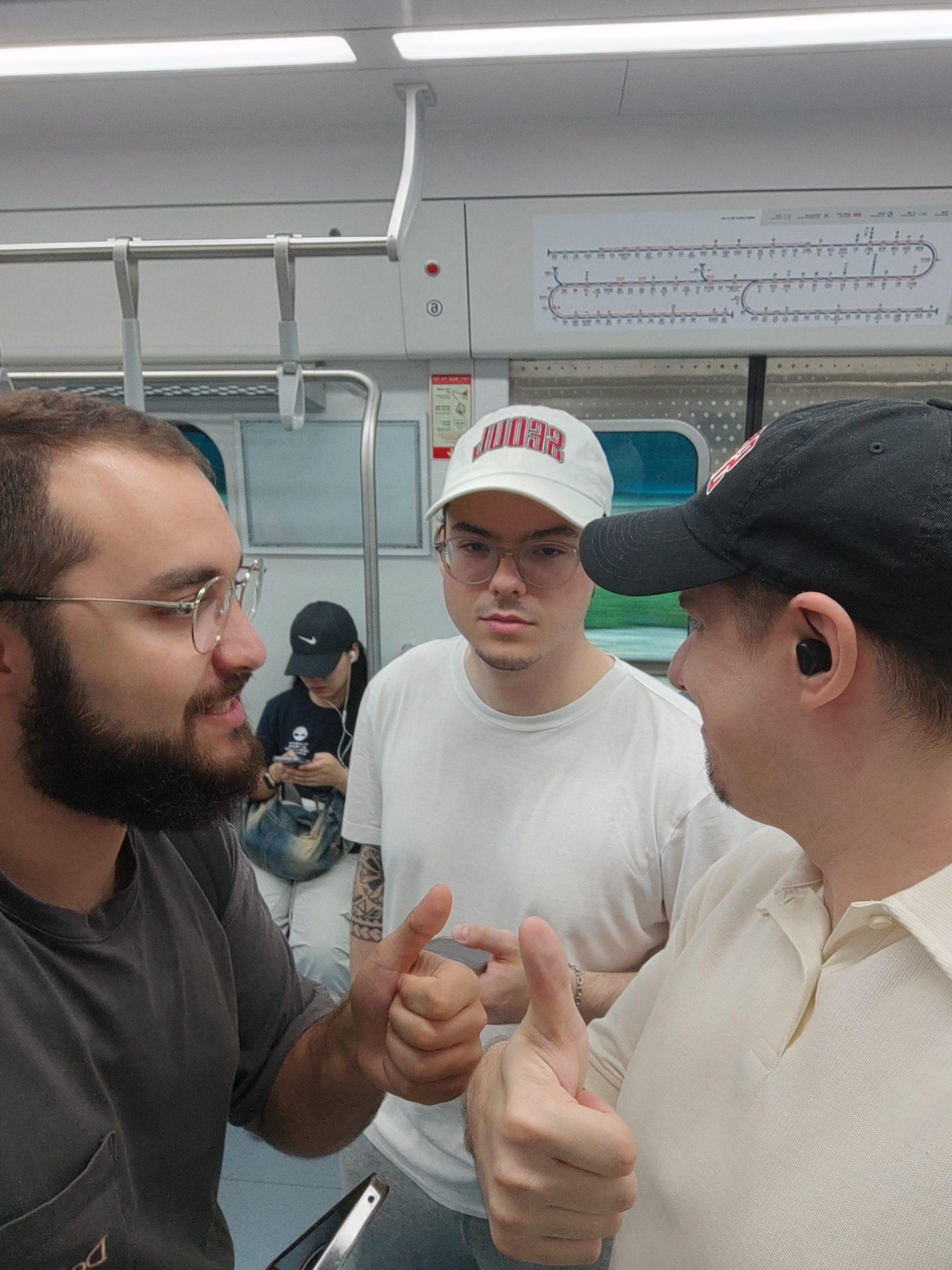 Drei Männer stehen in einem U-Bahn-Zug, zwei von ihnen zeigen den Daumen nach oben und lächeln. Eine weitere Person sitzt im Hintergrund und schaut auf ein Telefon. Hinter ihnen sind ein U-Bahn-Plan und ein Fenster mit verschwommener Landschaft zu sehen.