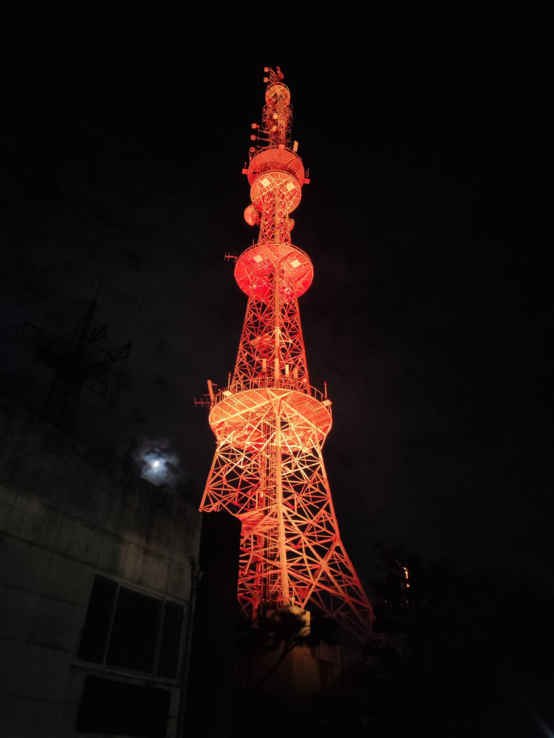 Ein hoher, orange-rot beleuchteter Sendeturm steht vor einem dunklen Nachthimmel, an dem einige Wolken und ein schwacher Mond im Hintergrund zu sehen sind. Nahegelegene Gebäude erscheinen im Schatten der Basis.