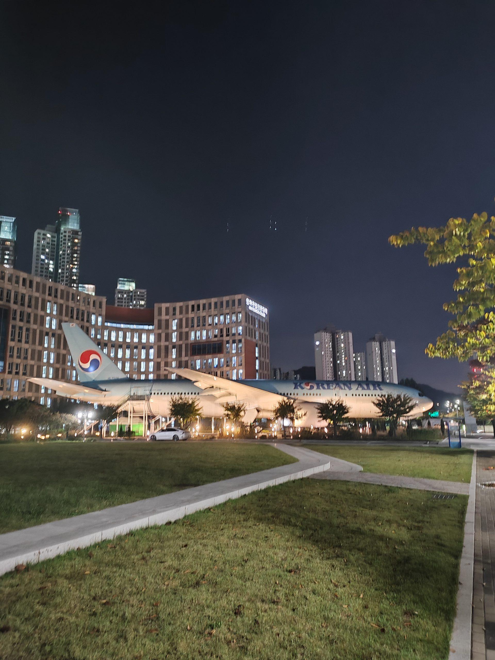 Ein Korean-Air-Flugzeug ist nachts im Freien zu sehen, umgeben von Gras, Bäumen und modernen Gebäuden mit beleuchteten Fenstern im Hintergrund.