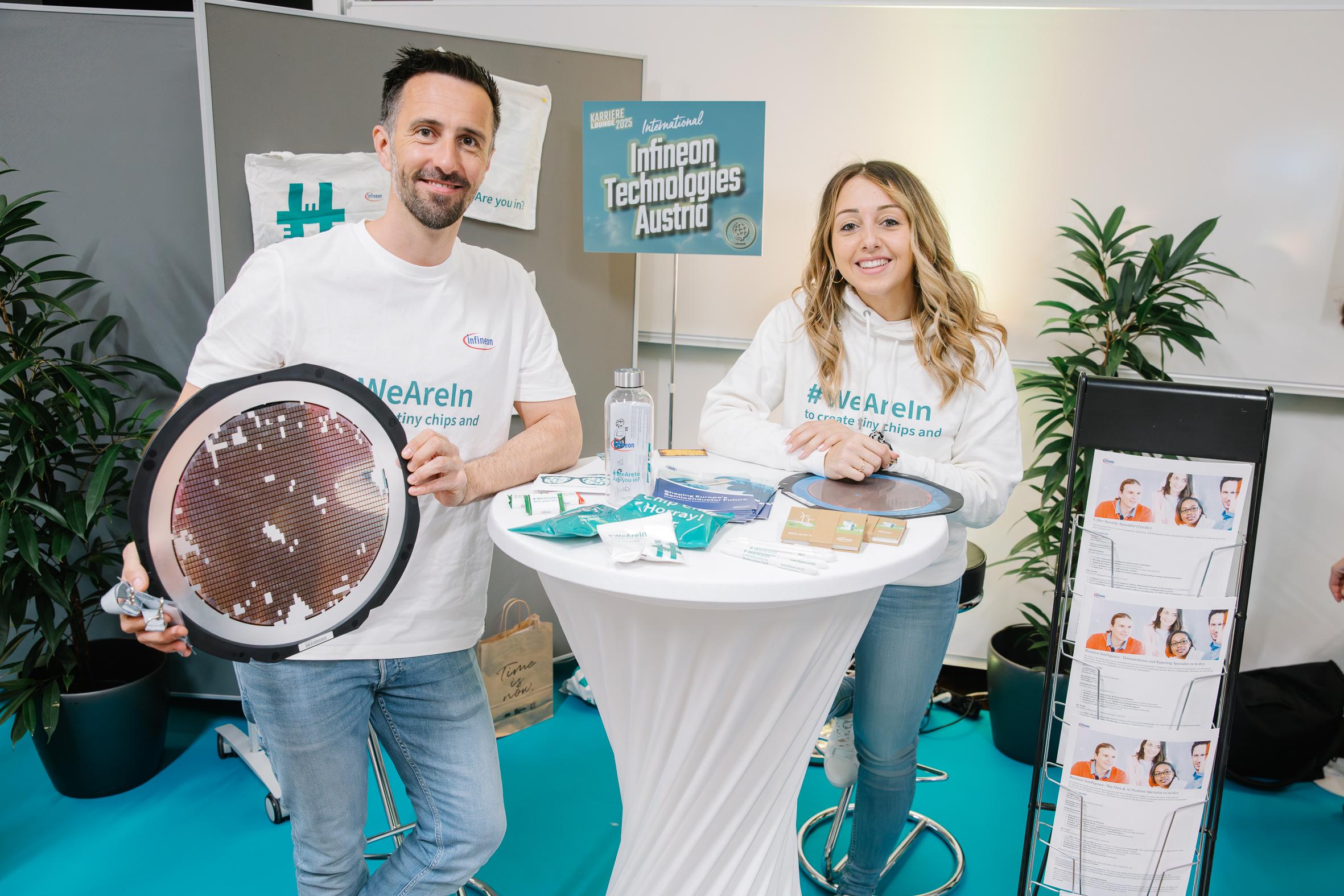 Zwei Personen in weißen #WeAreIn-Shirts stehen an einem Stand von Infineon Technologies Austria und legen technische Artikel und Broschüren aus. Sie lächeln in die Kamera, im Hintergrund sind Pflanzen und ein Schild zu sehen.