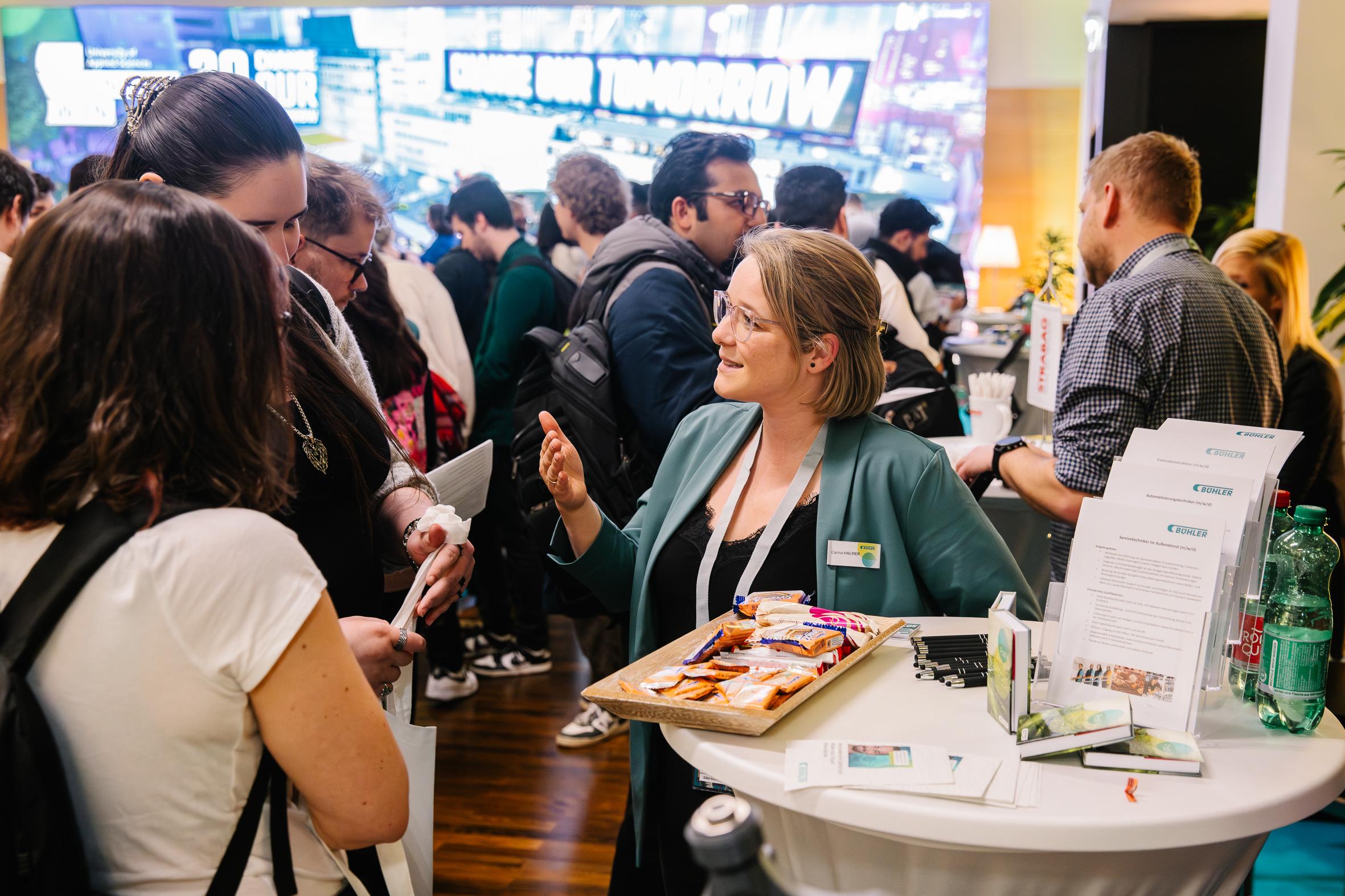 Eine Frau mit einem Namensschild spricht an einem belebten Konferenzstand mit den Teilnehmern und bietet Snacks von einem Tablett an. Die Leute versammeln sich um sie herum und unterhalten sich, während auf dem Tresen Broschüren und Getränke stehen.