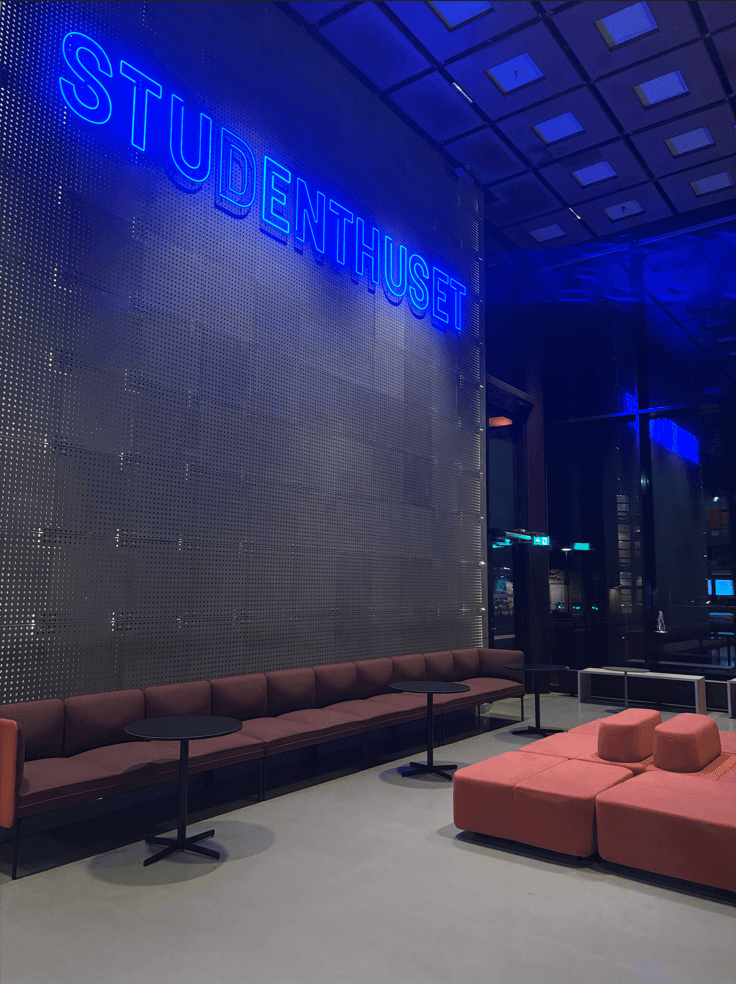 Modernes Interieur eines Gebäudes mit einem großen blauen STUDENTHUSET-Neonschild an einer perforierten Metallwand, mit kastanienbraunen Sitzmöbeln, rosa Hockern, kleinen Tischen und großen Glasfenstern.