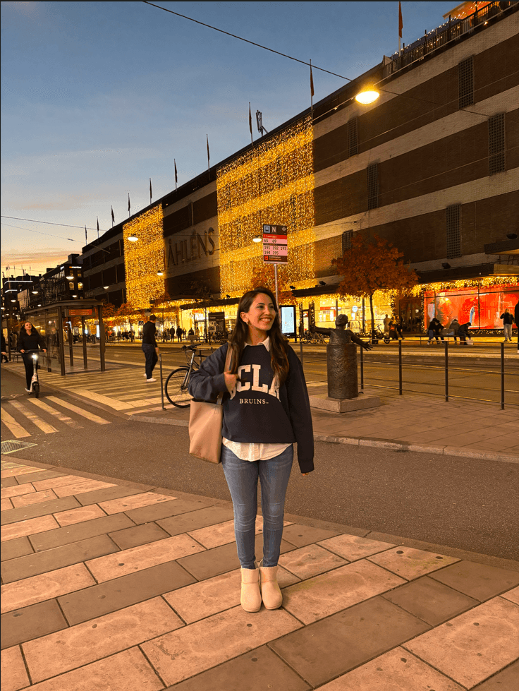 Eine Frau steht in der Abenddämmerung auf einem Bürgersteig und lächelt. Sie trägt ein dunkles UCLA-Sweatshirt, Jeans und Stiefel und hat eine beige Tasche dabei. Hinter ihr ist das Kaufhaus Åhléns mit festlichen Lichtern beleuchtet. Im Hintergrund sind andere Menschen zu sehen.