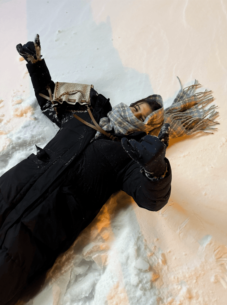 Eine Person in schwarzem Mantel, Schal und Handschuhen liegt auf dem Rücken im Schnee, lächelt und macht mit beiden Händen die Geste des "Rock on". Eine kleine Handtasche liegt quer über ihrem Körper.