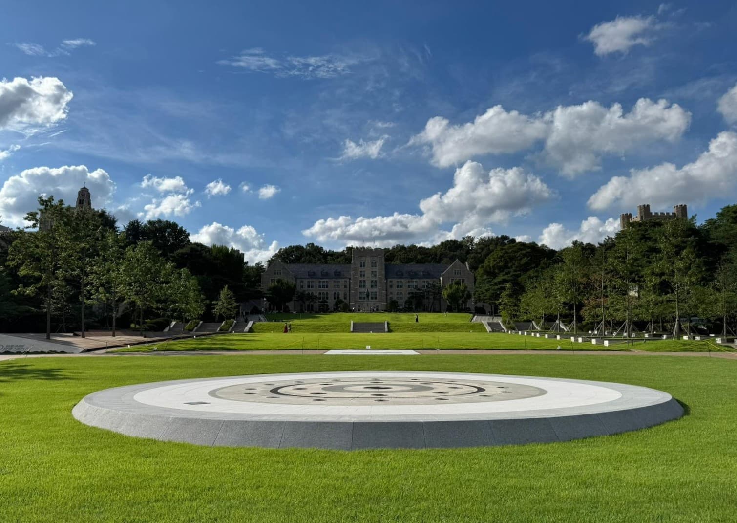 Ein großer kreisförmiger Stein liegt in der Mitte eines grünen Rasens, umgeben von Bäumen und Wegen, mit einem schlossähnlichen Gebäude und einem blauen Himmel mit Wolken im Hintergrund.