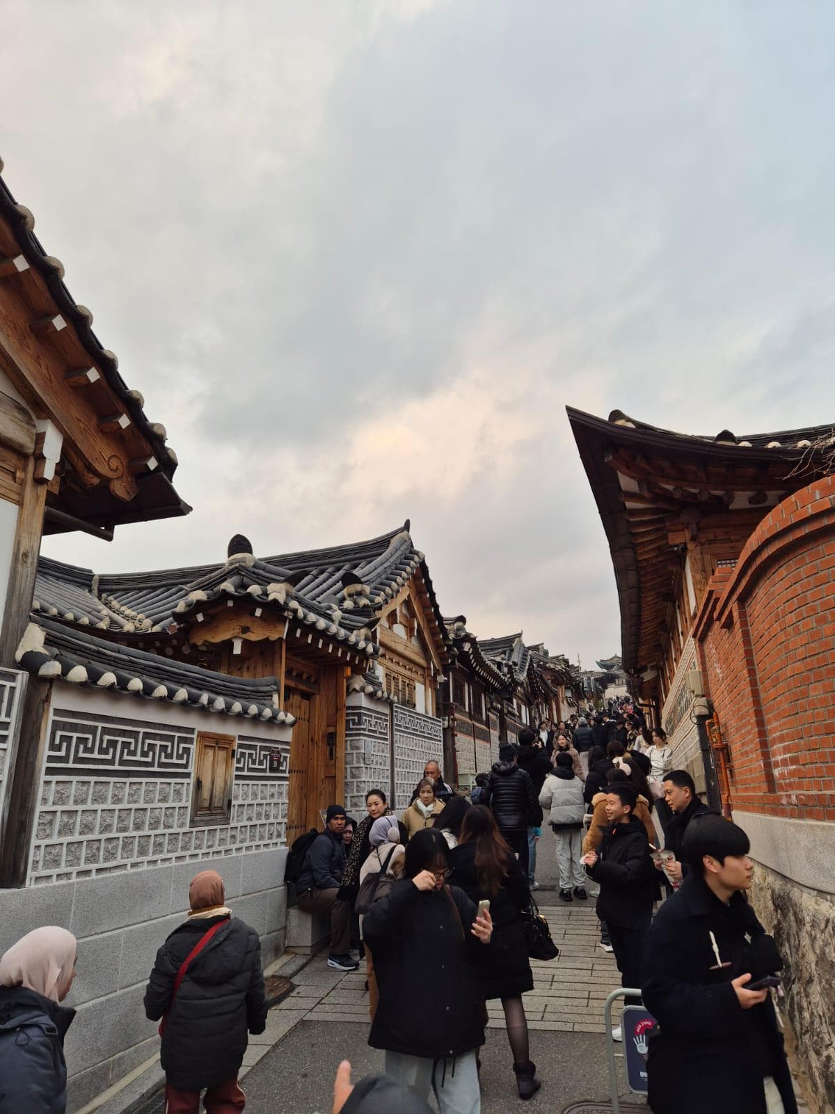 Eine belebte Straßenszene mit vielen Menschen, die zwischen traditionellen koreanischen Hanok-Häusern bei bewölktem Himmel spazieren gehen und Fotos machen. Die Häuser haben Ziegeldächer und gemusterte Wände, die ein historisches Ambiente schaffen.