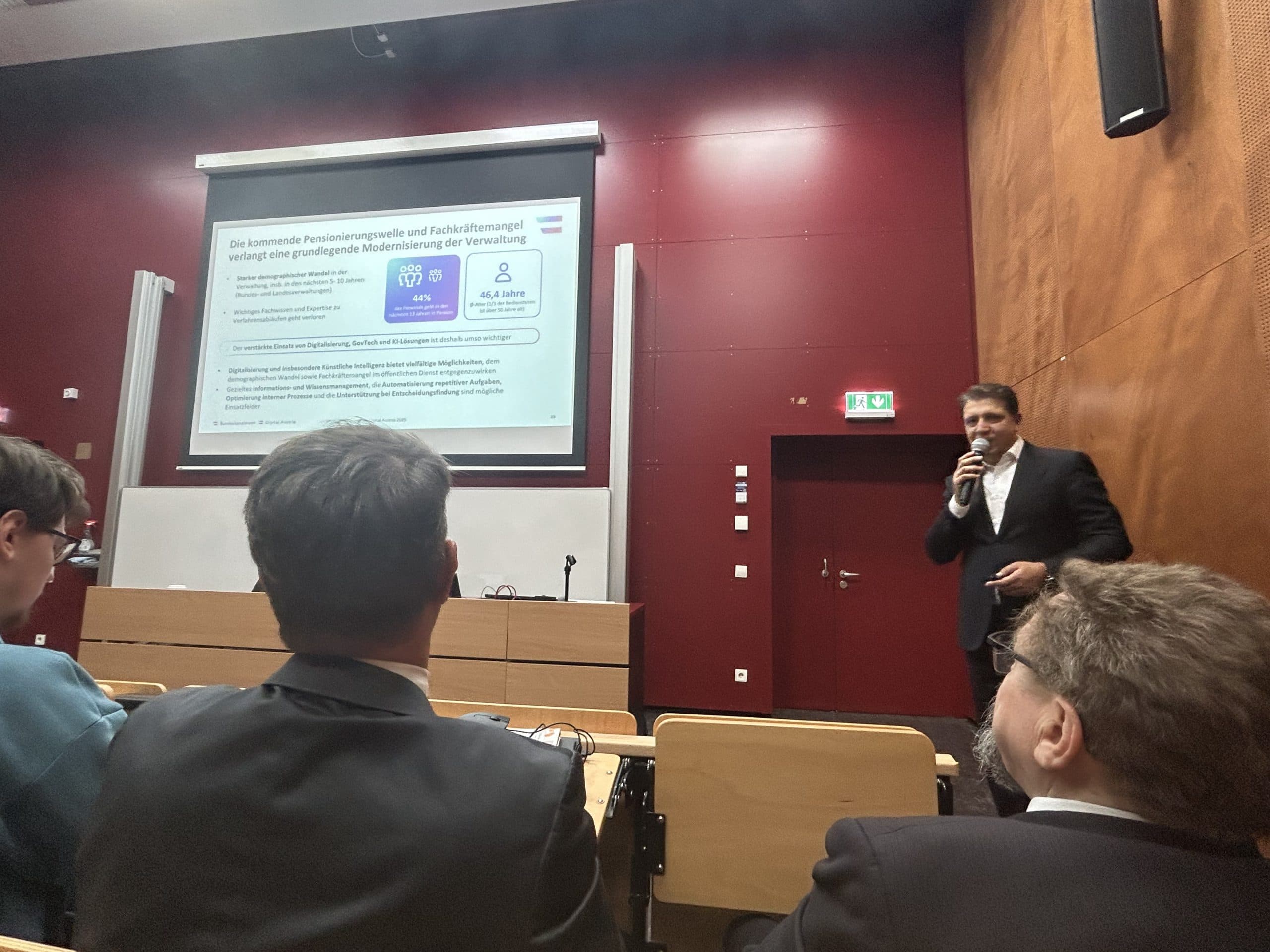 Ein Mann im Anzug spricht in einem Hörsaal in ein Mikrofon und präsentiert dem Publikum eine Folie in deutscher Sprache. Mehrere Personen sitzen ihm gegenüber, der Raum hat rote Wände und eine Holzverkleidung.