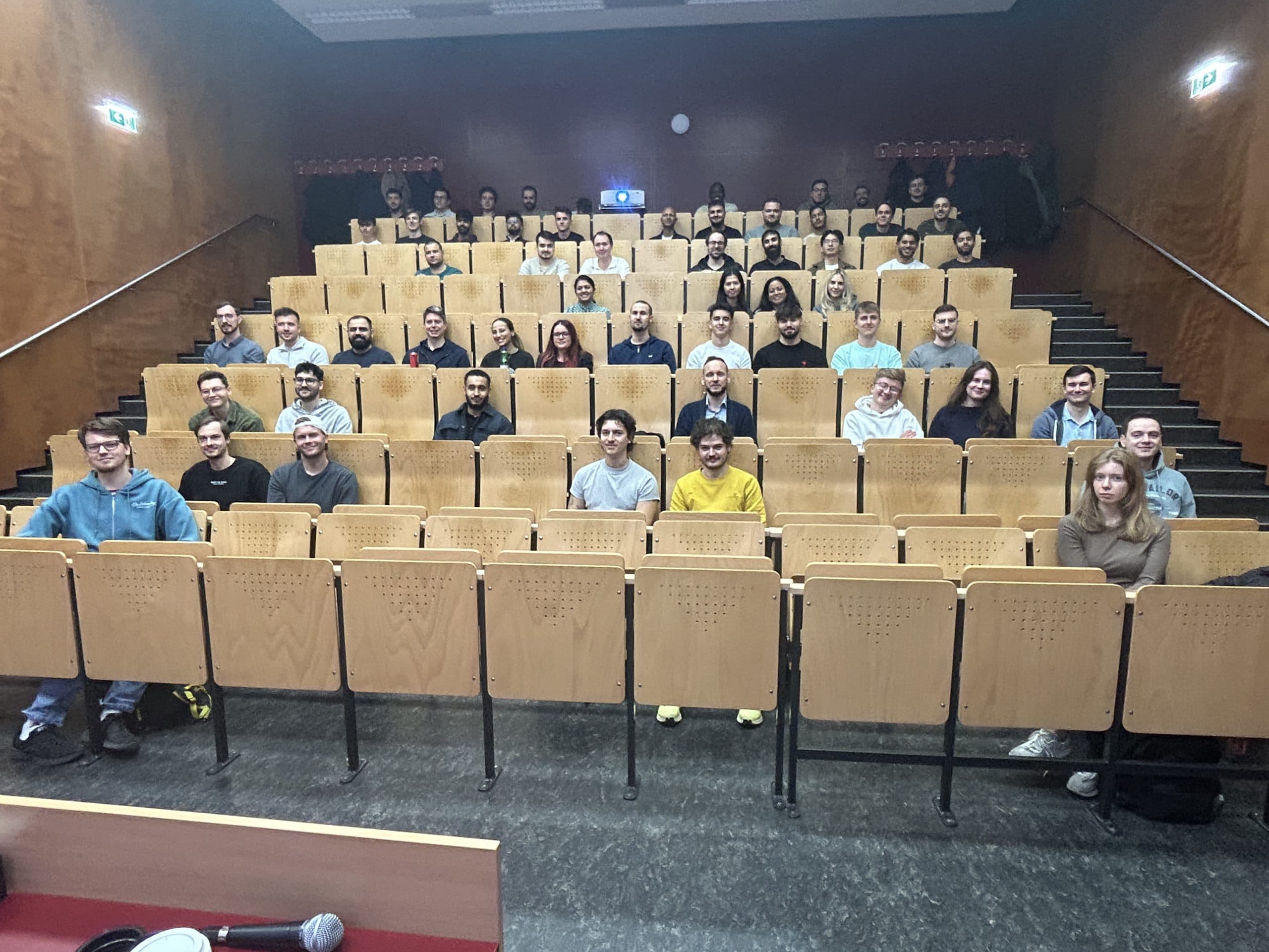 Eine Gruppe von Menschen, zumeist Erwachsene, sitzt in mehreren Reihen hölzerner Hörsaalstühle in einem Vortragssaal und blickt in die Kamera. Der Raum hat braune Wände und eine Oberlichtbeleuchtung.