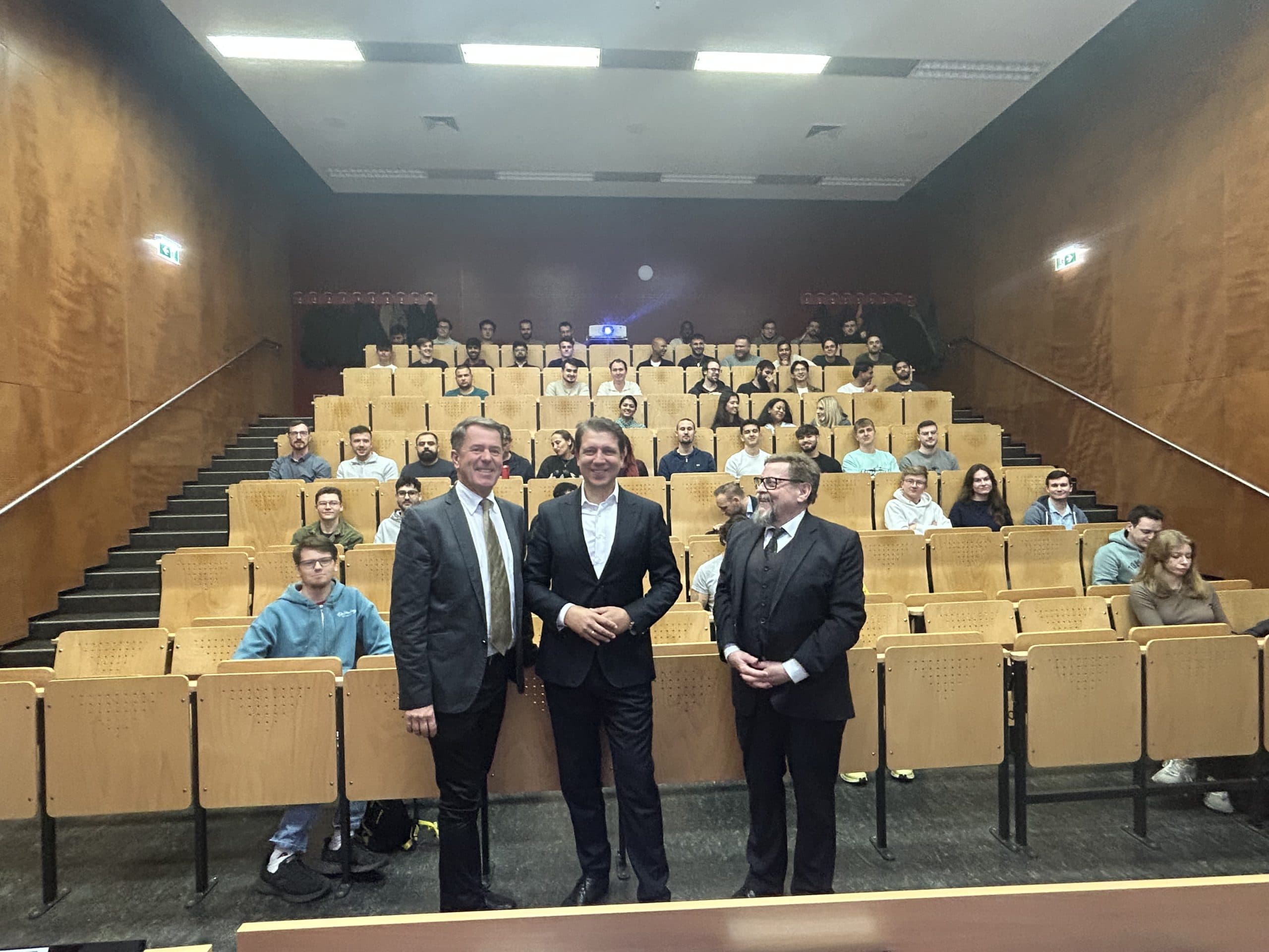 Drei Männer in Anzügen stehen am Anfang eines Hörsaals, der mit sitzenden Studenten gefüllt ist. Das Klassenzimmer hat Holzwände und eine Reihenbestuhlung, die Leute lächeln und schauen in die Kamera.