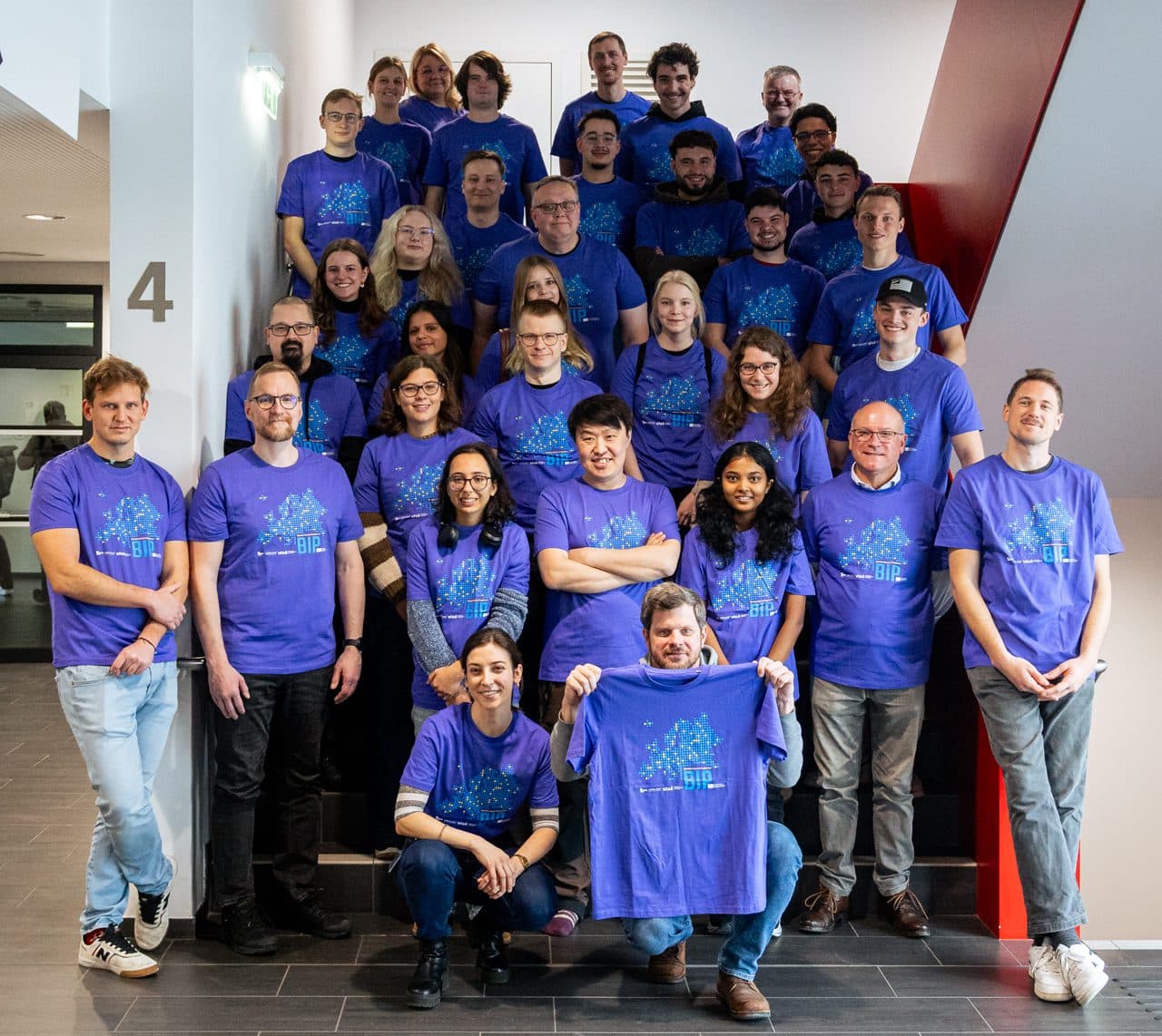 Eine Gruppe von Personen, die zusammenpassende lila T-Shirts tragen, posieren gemeinsam auf einer Treppe in einem Innenraum und lächeln in die Kamera. Die Gruppe scheint sowohl aus Männern als auch aus Frauen unterschiedlichen Alters zu bestehen.
