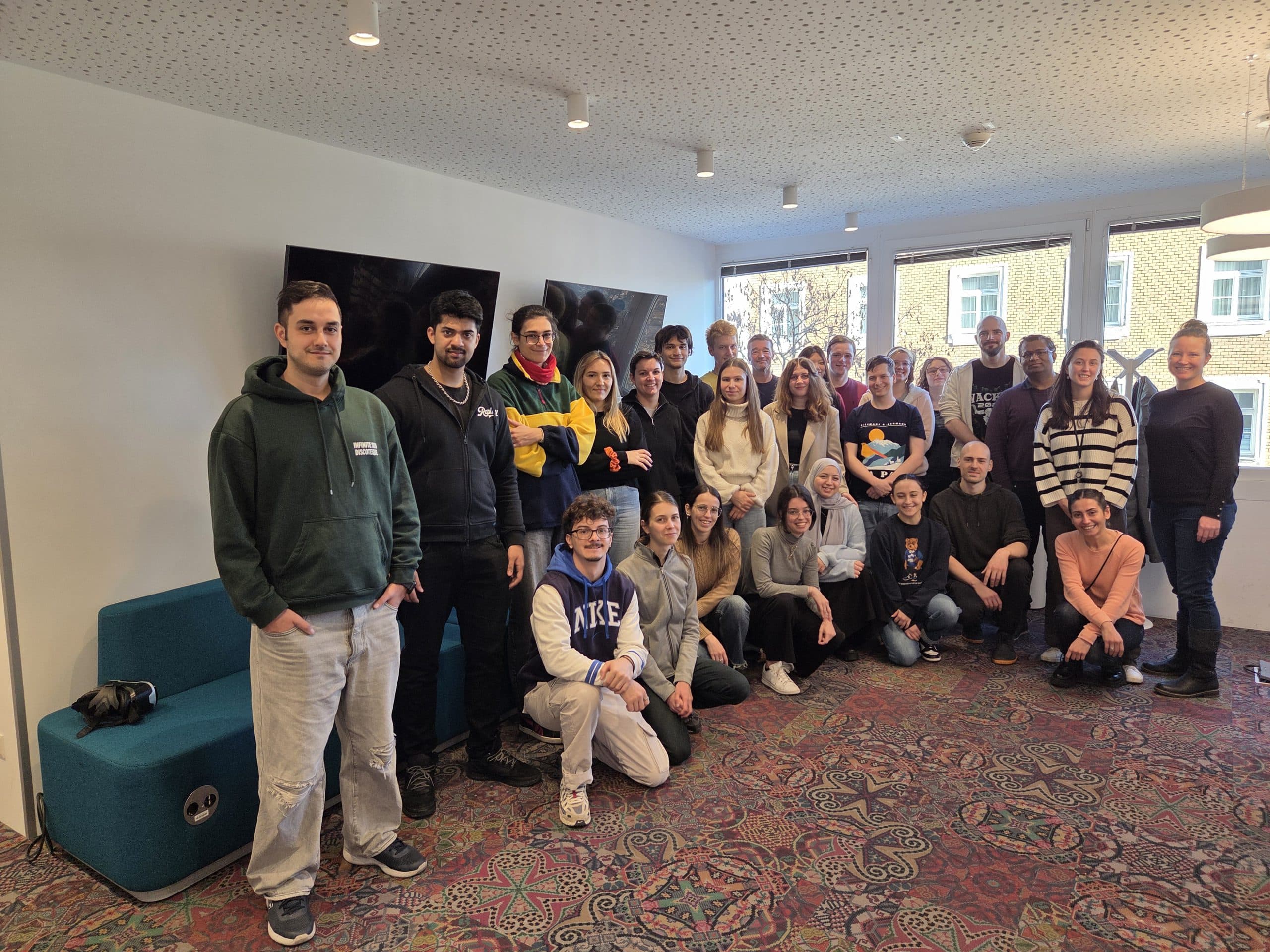 Eine Gruppe von etwa 30 Personen, die in einem Innenraum in ungezwungener Atmosphäre zusammenstehen oder -sitzen und in die Kamera lächeln. Im Hintergrund sind große Fenster, ein Fernseher und ein gemusterter Teppich zu sehen.