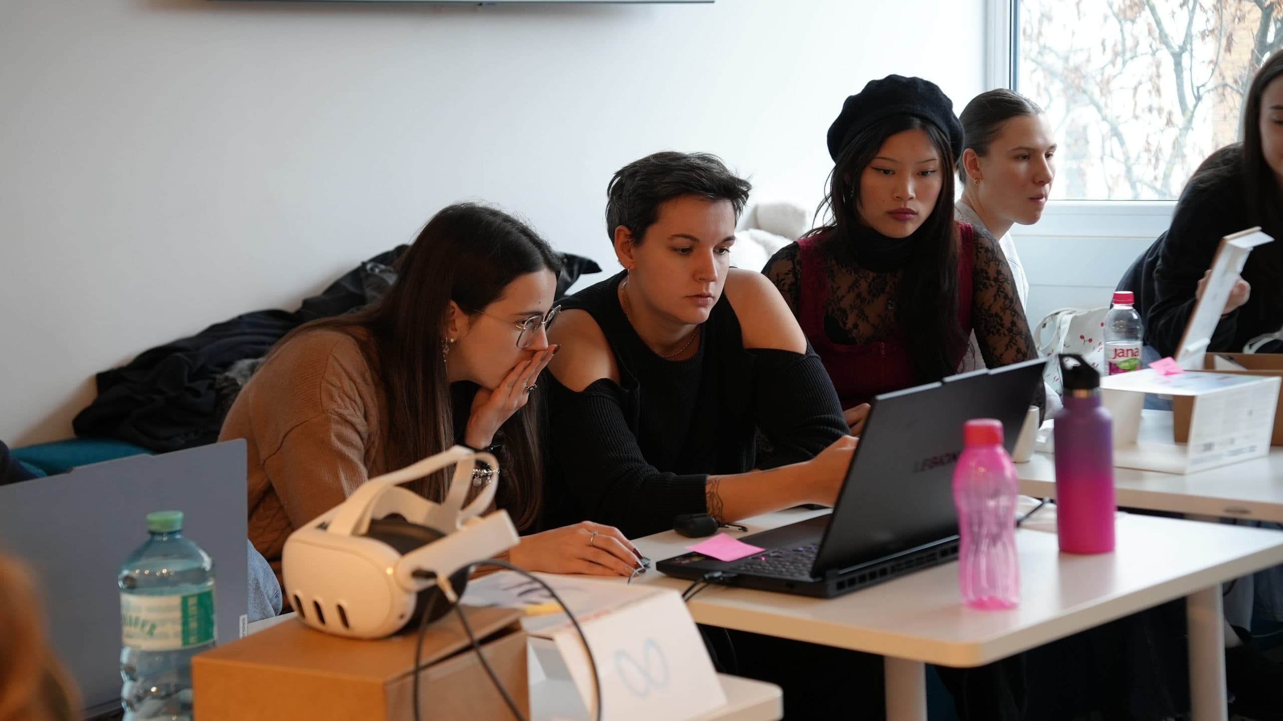 Eine Gruppe konzentrierter junger Erwachsener sitzt an einem Tisch mit Laptops, Notebooks und Wasserflaschen und arbeitet in einem hellen, modernen Raum zusammen. Im Vordergrund sind ein VR-Headset und ein Pappkarton zu sehen.