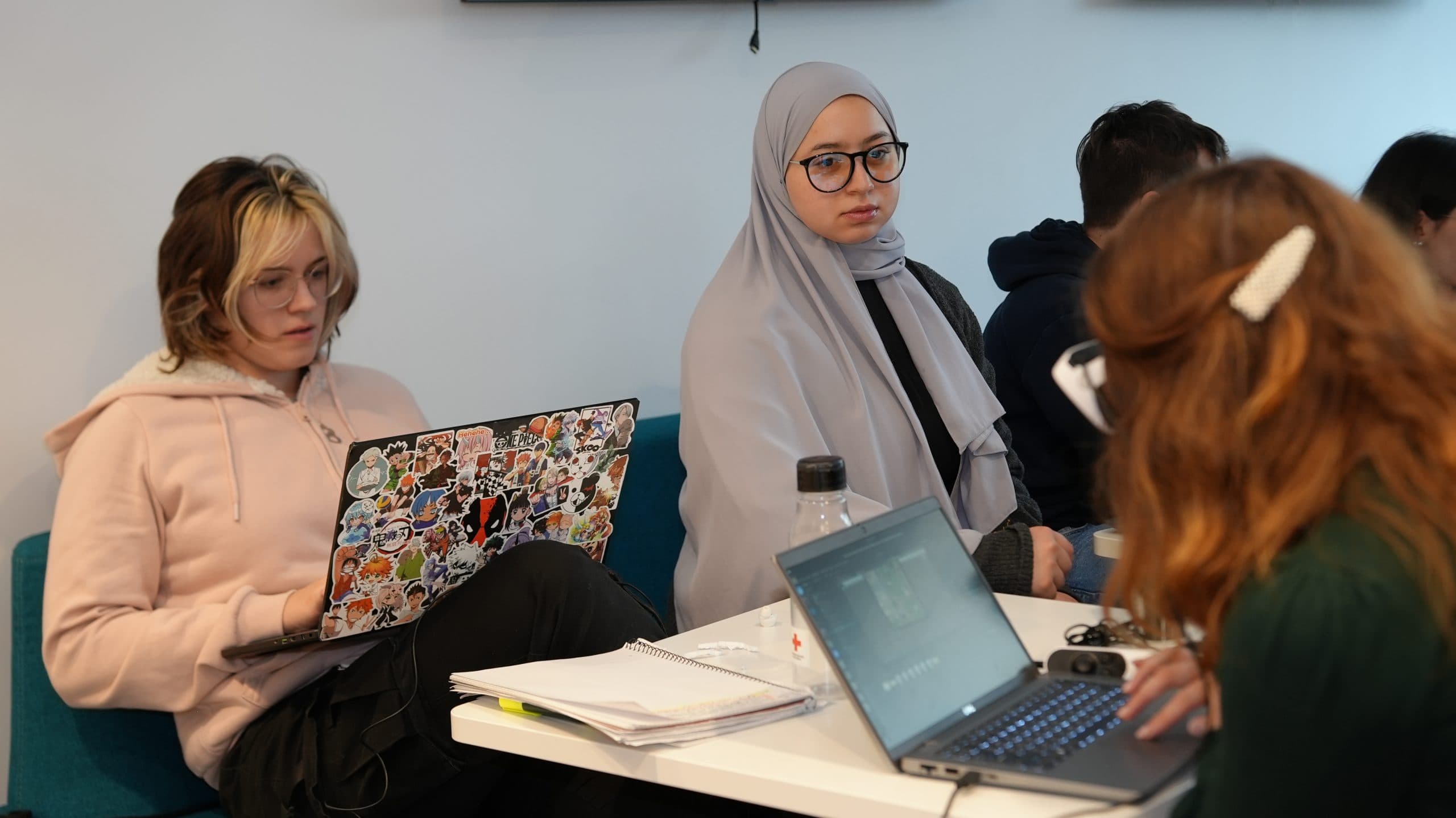 Drei Schüler sitzen an einem Tisch mit Laptops, Notizbüchern und einer Wasserflasche. Eine Schülerin trägt einen Hidschab und eine Brille, eine hat blonde Haare und einen Laptop mit Aufklebern, und eine andere schaut mit einer Haarspange weg.