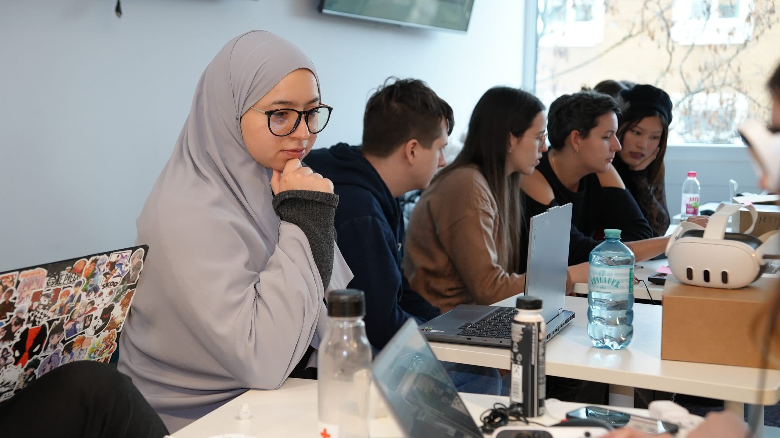 Eine Frau mit hellgrauem Hidschab und Brille sitzt an einem Tisch mit anderen jungen Erwachsenen, die alle in einem hellen, modernen Klassenzimmer mit Laptops arbeiten. Wasserflaschen und elektronische Geräte sind auf dem Tisch verstreut.