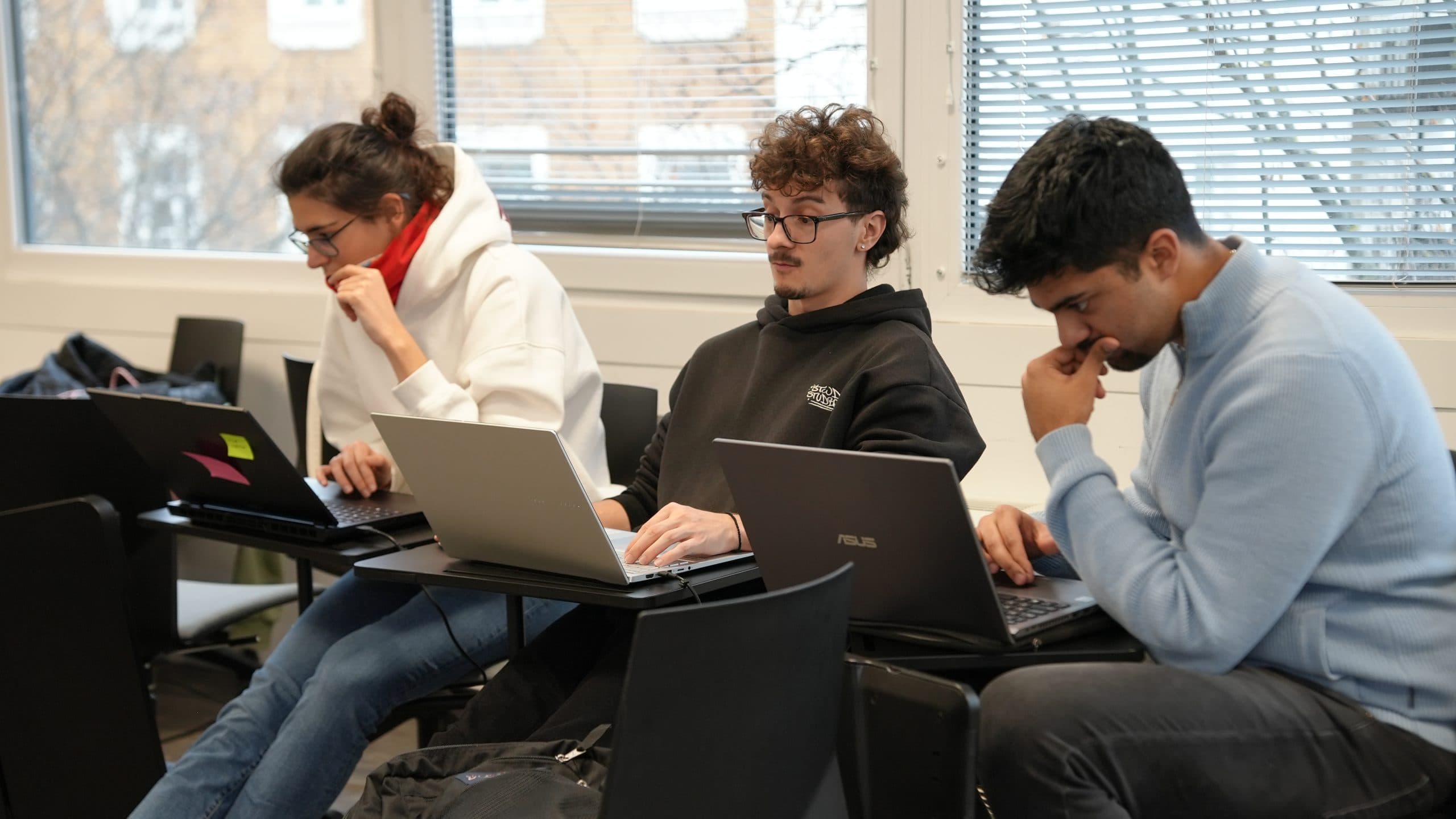 Drei Personen sitzen in einem Klassenzimmer, alle auf ihre Laptops konzentriert. Sie scheinen zu arbeiten oder zu lernen, mit einem Fenster und Jalousien im Hintergrund.