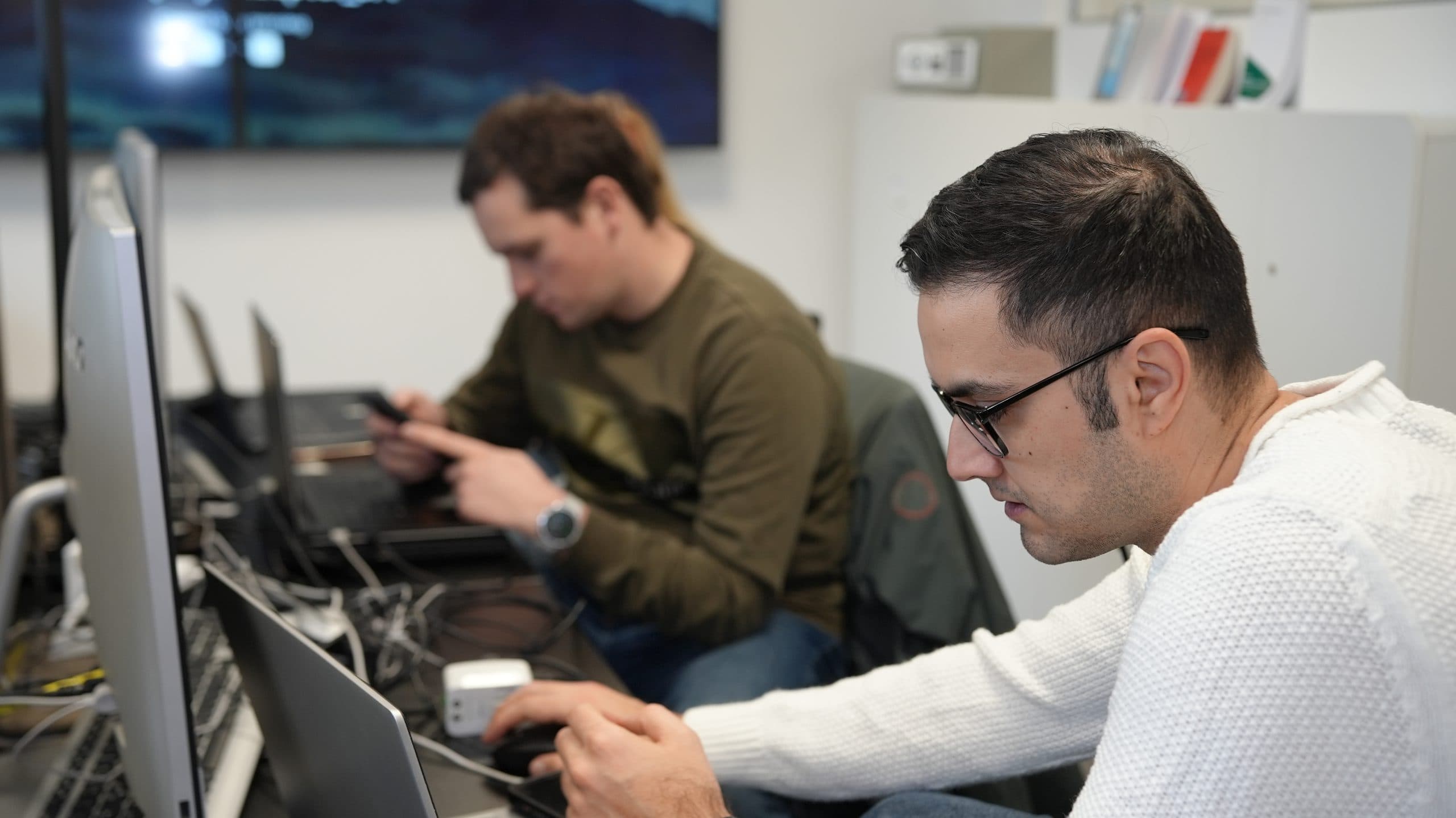 Zwei Männer sitzen an Schreibtischen und arbeiten an Laptops in einer Büroumgebung. Der Mann im Vordergrund trägt eine Brille und einen weißen Pullover, während der Mann im Hintergrund ein grünes Hemd trägt und auf sein Telefon konzentriert ist.