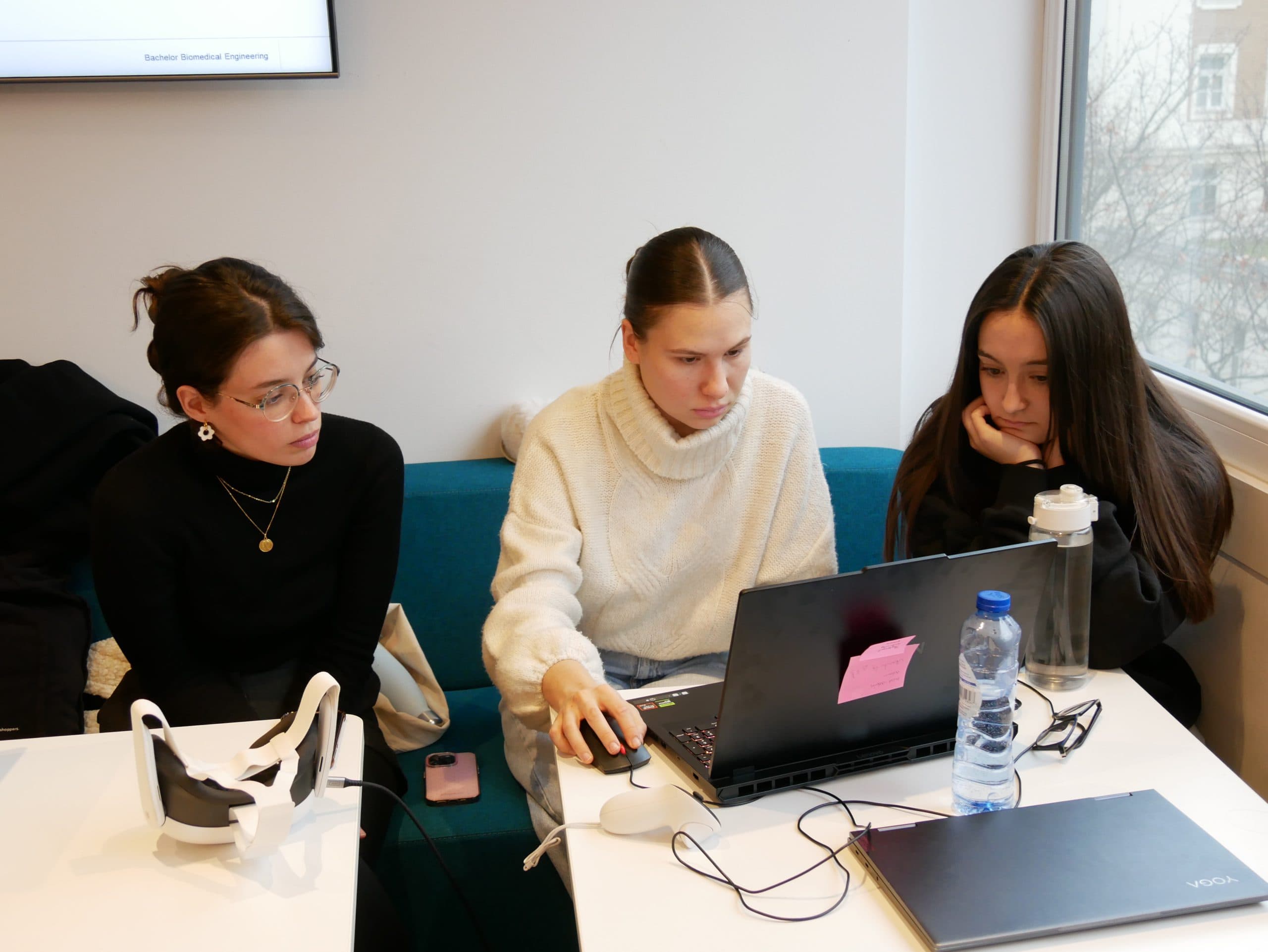 Drei junge Frauen sitzen zusammen an einem Tisch und arbeiten konzentriert an einem Laptop. Eine bedient die Maus, während die anderen zusehen. Ein Virtual-Reality-Headset, eine Wasserflasche und ein Telefon liegen auf dem Tisch. Im Hintergrund ist ein Fenster zu sehen.