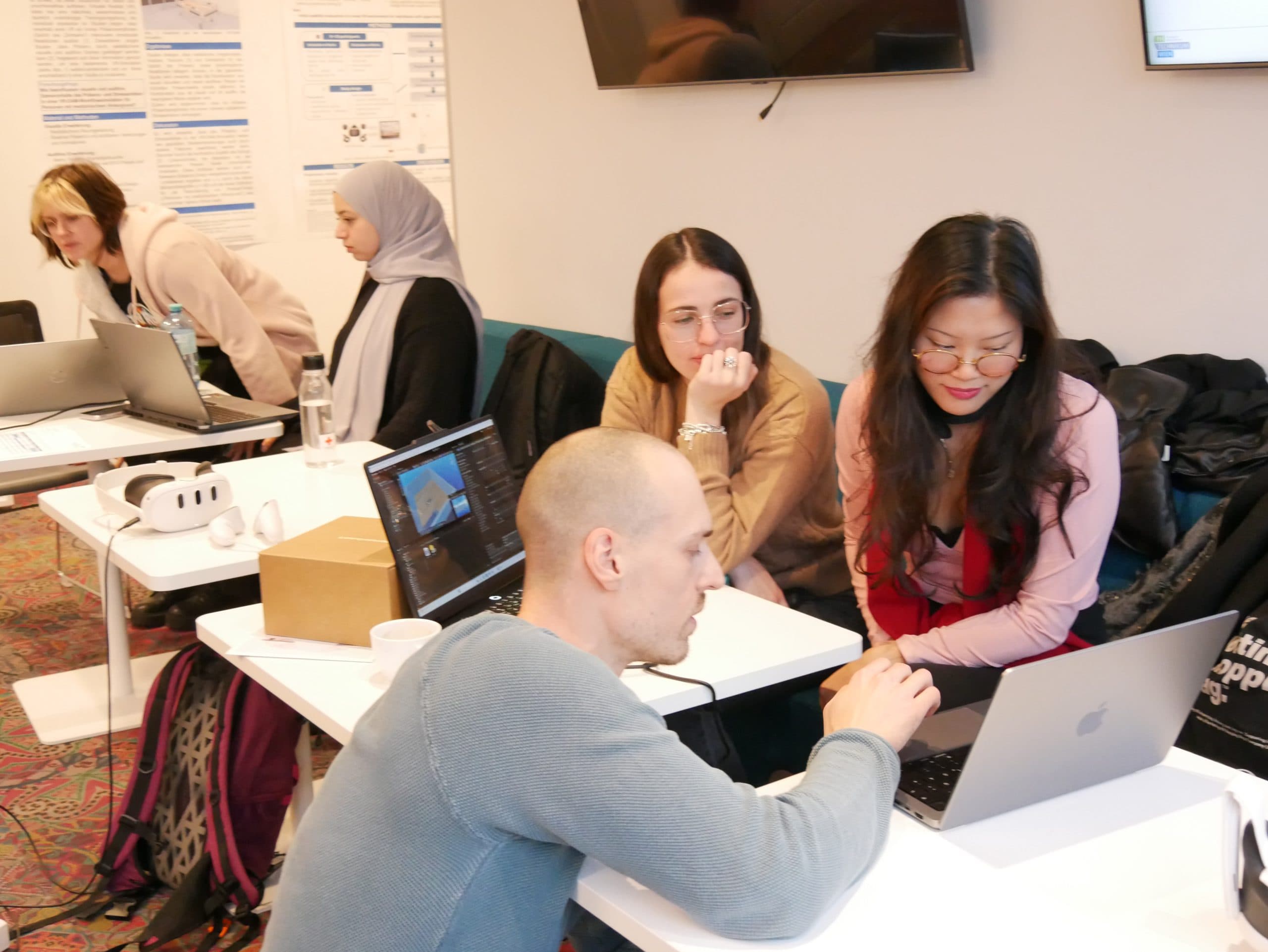 Eine Gruppe von Personen sitzt an Tischen und arbeitet in einem Klassenzimmer an Laptops. Zwei Frauen beobachten, wie ein Mann auf einen Laptop-Bildschirm zeigt. Im Hintergrund sind andere Personen ebenfalls mit Diskussionen und Computerarbeit beschäftigt.