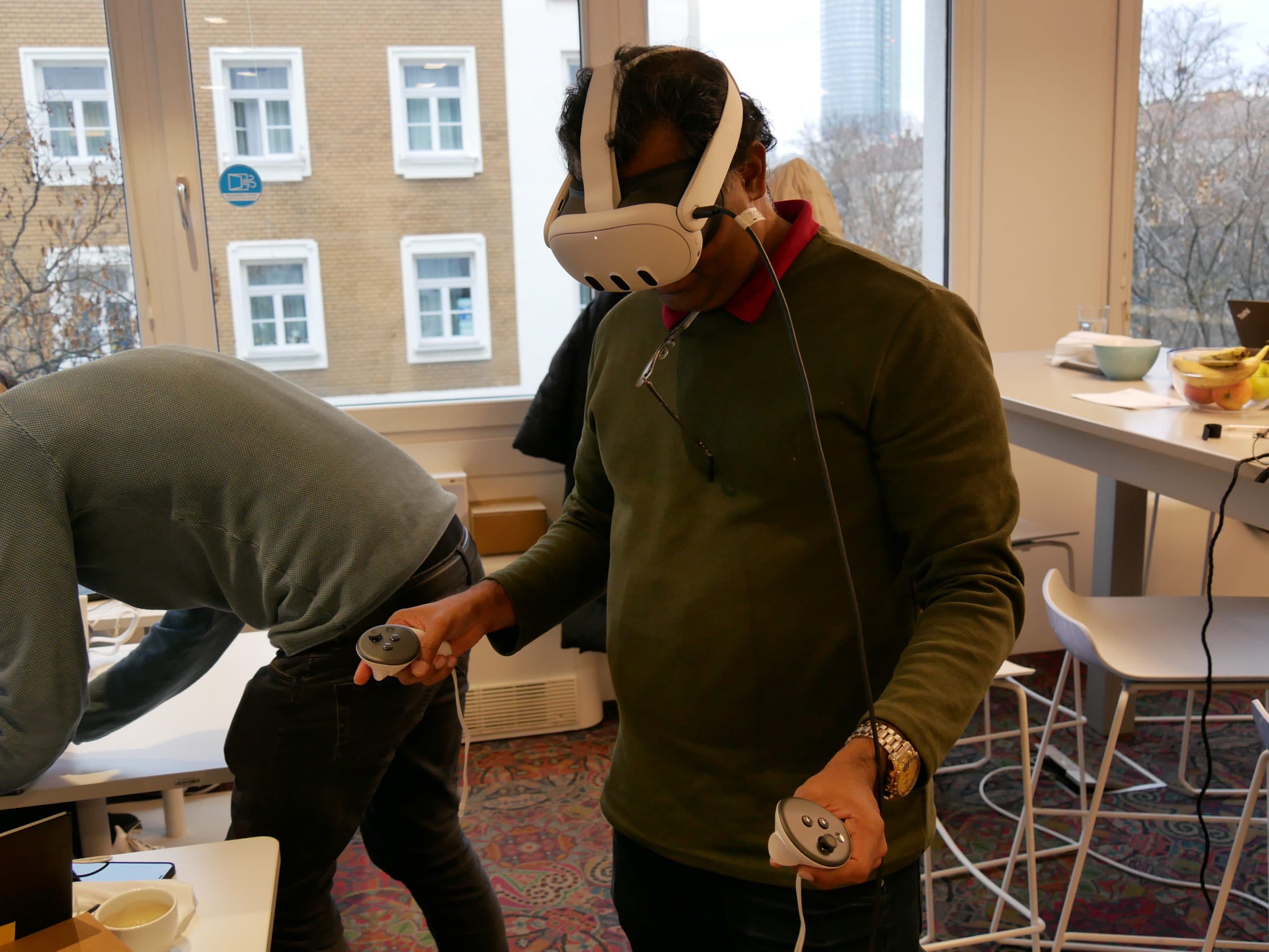 Eine Person mit einem Virtual-Reality-Headset und Controllern steht in einem Raum mit großen Fenstern. Eine weitere Person ist über einen Tisch in der Nähe gebeugt. Im Raum sind Hocker, Tische und einige Lebensmittel zu sehen.