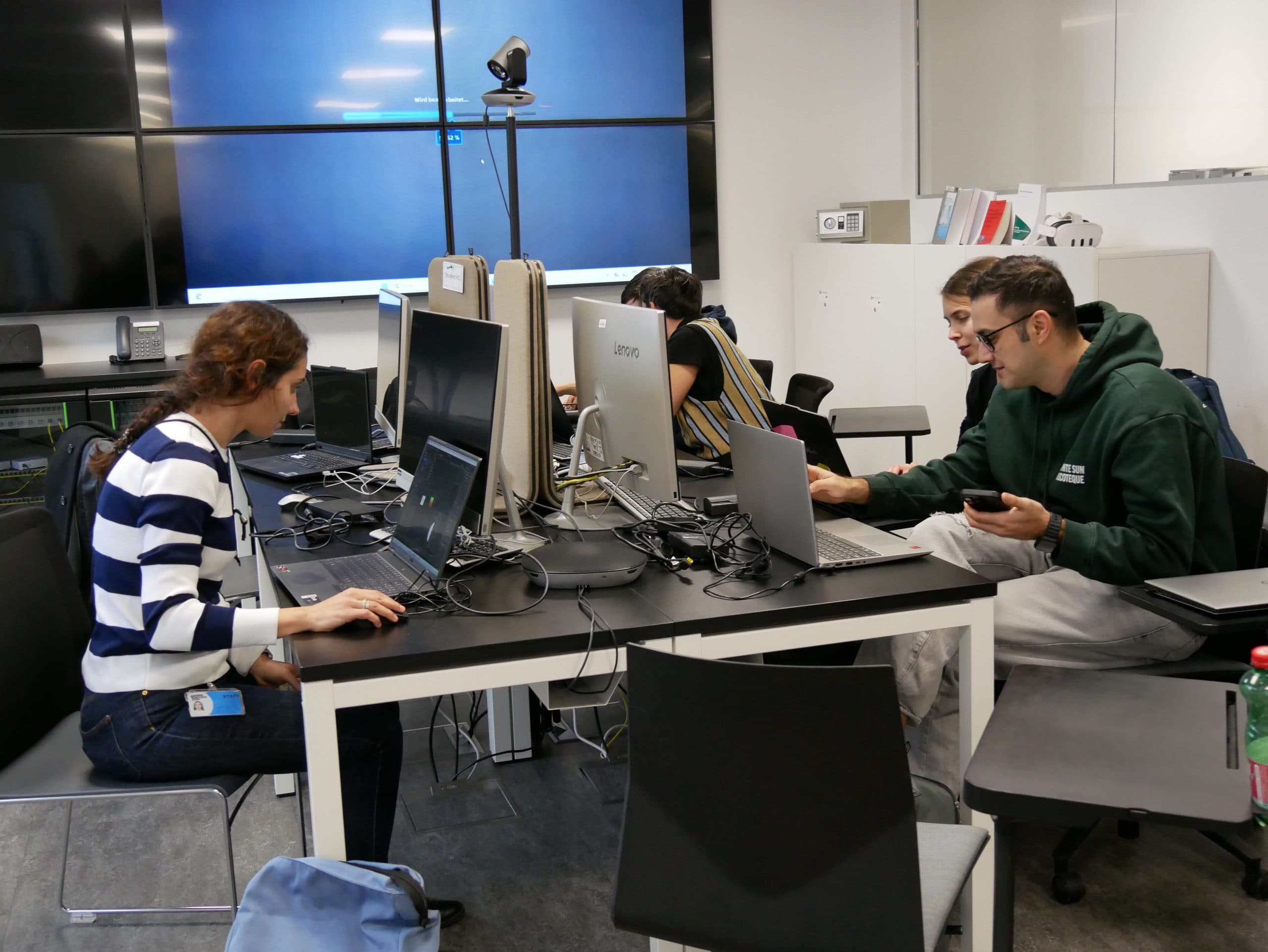 Vier Personen sitzen an einem Tisch und arbeiten an Laptops in einem modernen Büro oder Klassenzimmer. Der Raum ist mit großen Monitoren an der Wand, verschiedenen elektronischen Geräten und verstreuten Stühlen und Taschen ausgestattet.