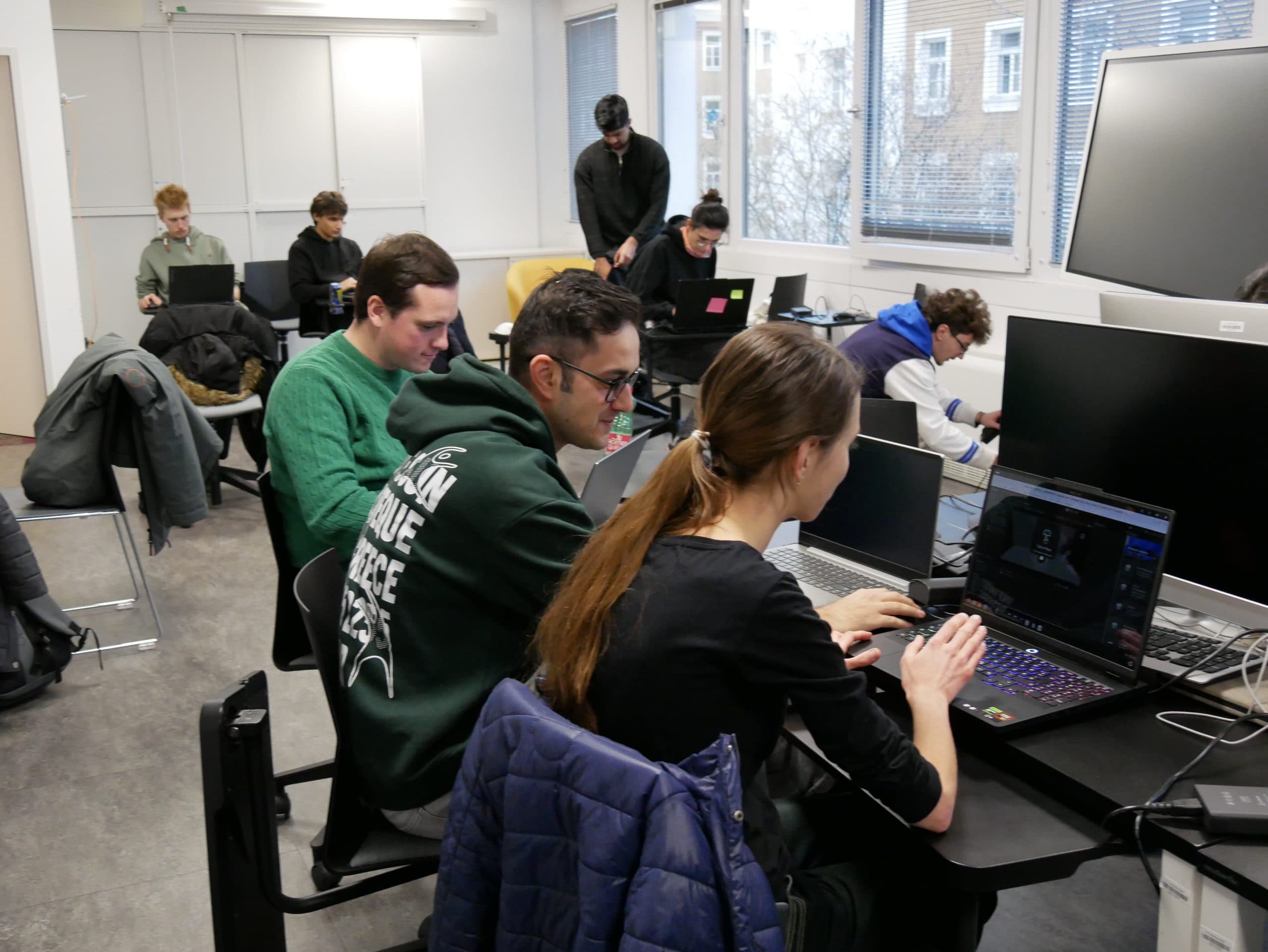 In einem modernen Klassenzimmer mit großen Fenstern sitzen mehrere Personen und arbeiten an Laptops. Einige arbeiten zu zweit, andere allein. Es gibt Jacken auf den Stühlen und einen großen Bildschirm an der Stirnseite.