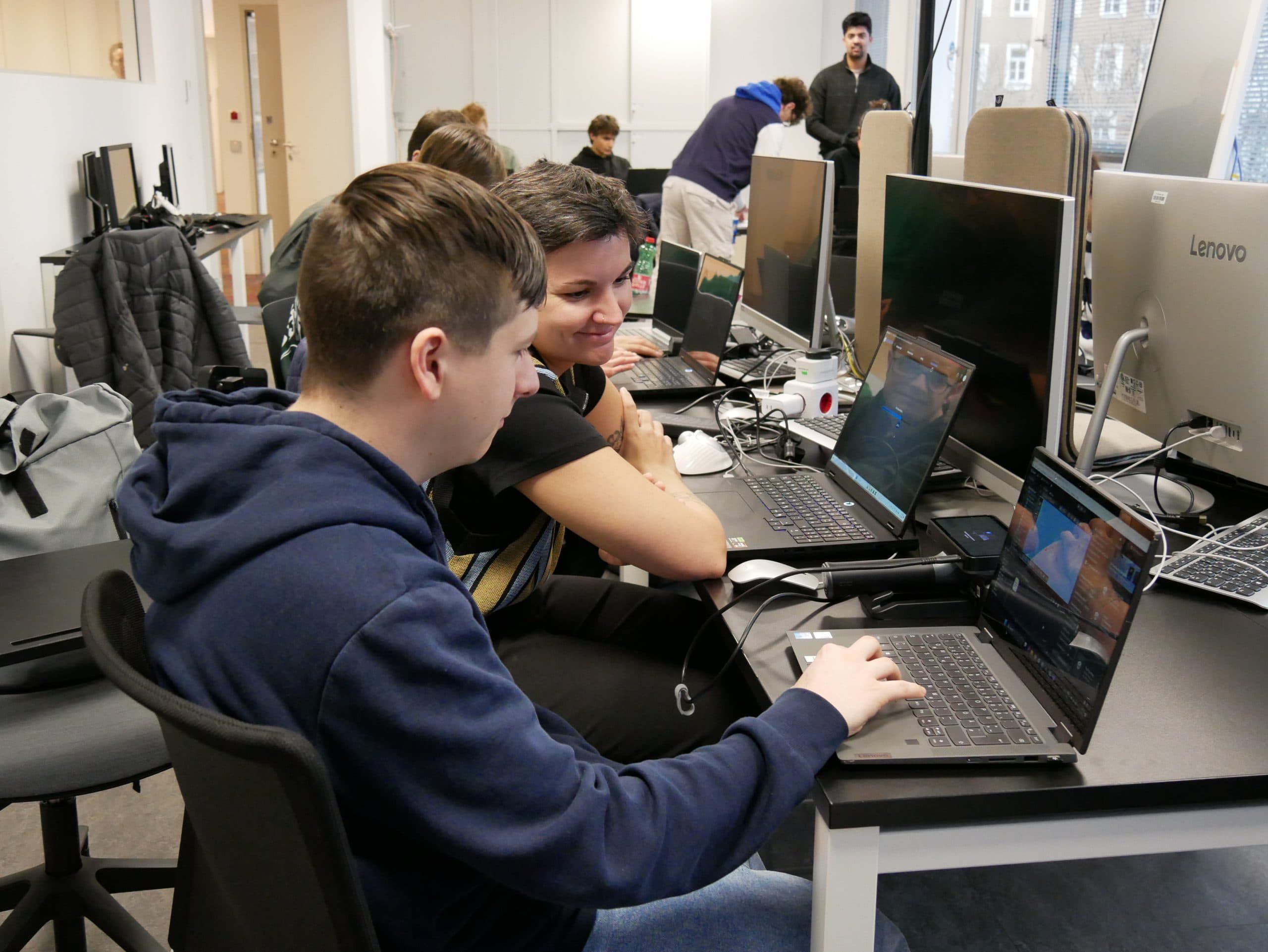 Ein Jugendlicher und ein Erwachsener sitzen Seite an Seite an einem Schreibtisch, beide konzentriert auf einen Laptop-Bildschirm. Im Hintergrund arbeiten mehrere andere Personen an Computern in einem modernen Klassenzimmer oder Computerraum.