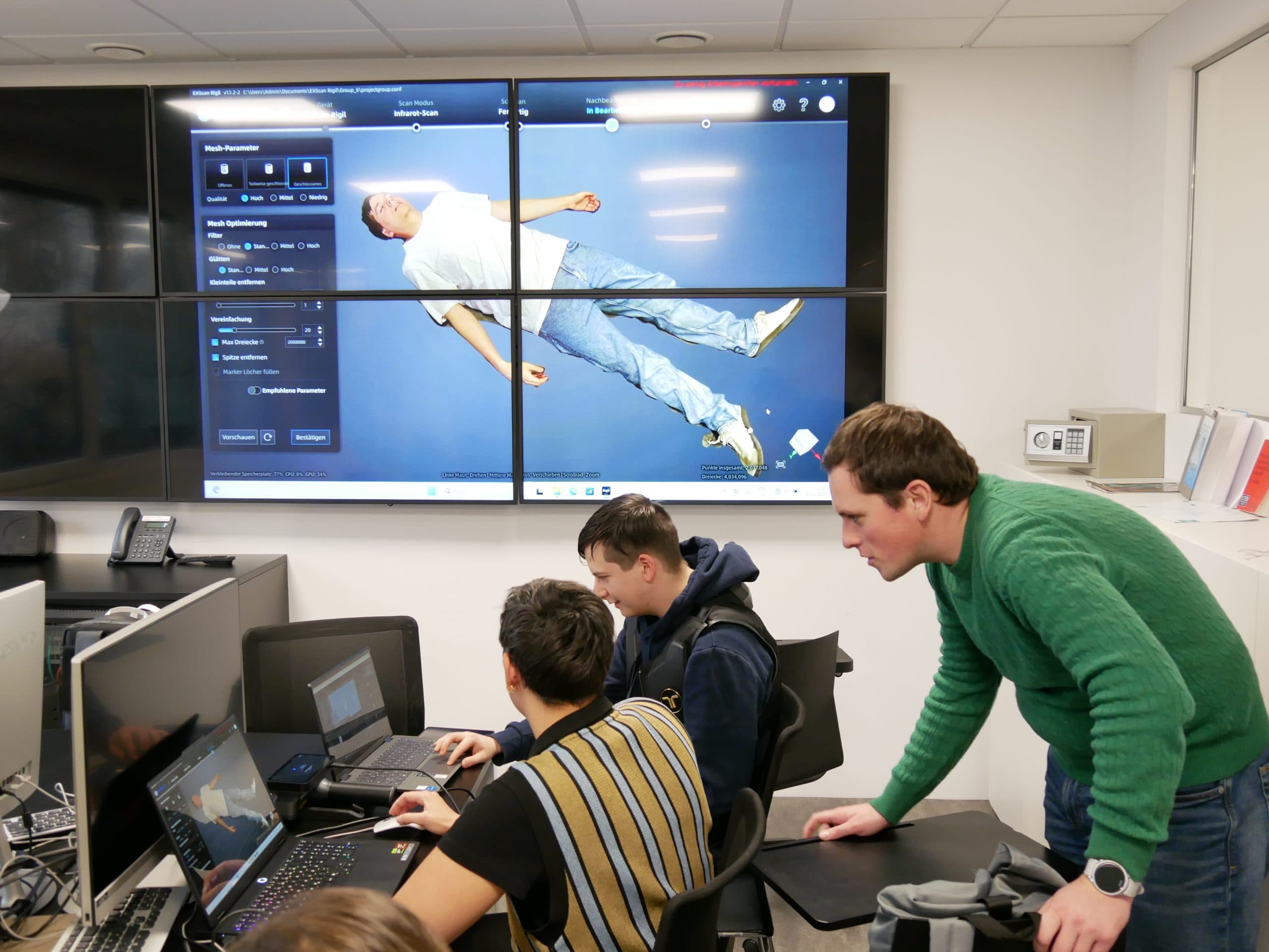 Drei Personen arbeiten in einem Büro an Computern, während ein Mann in einem grünen Pullover auf die Bildschirme schaut. Auf einem großen Wandmonitor ist das Bild einer liegenden Person zu sehen, auf dem eine Software-Schnittstelle zu sehen ist.