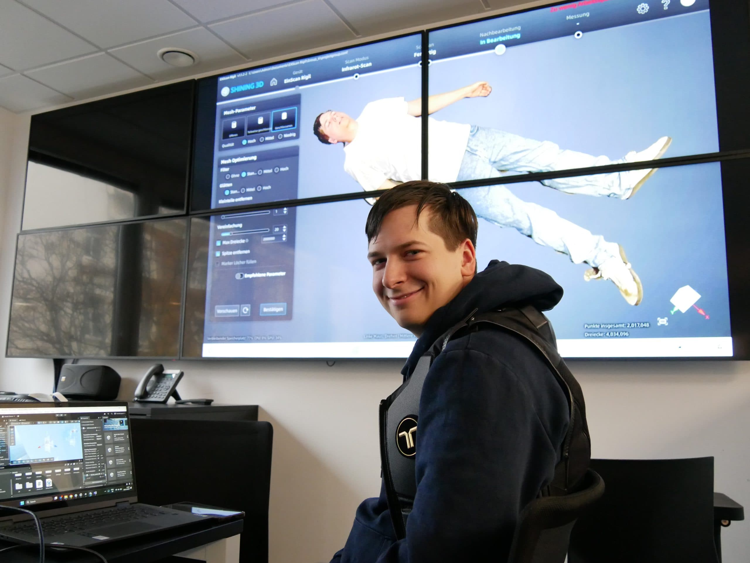 Ein Mann in einer Weste sitzt an einem Schreibtisch mit einem Laptop und lächelt in die Kamera. Hinter ihm zeigt ein großer Bildschirm das Bild einer liegenden Person und eine Benutzeroberfläche mit Bedienelementen und Daten.