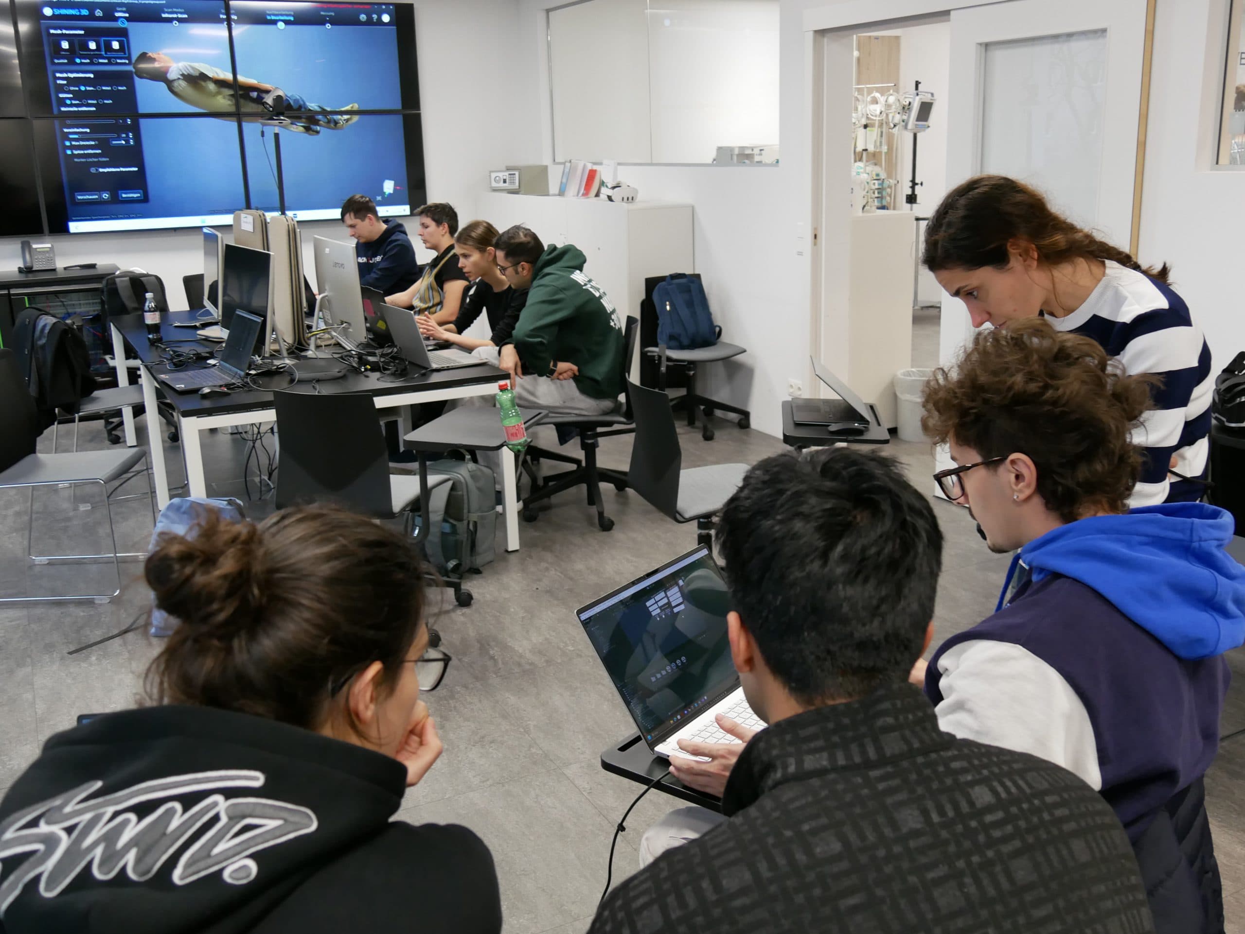 Schüler sitzen an Schreibtischen mit Computern in einem Klassenzimmer und arbeiten an digitalen Projekten. Ein Lehrer hilft einer Gruppe im Vordergrund, während im Hintergrund ein 3D-Modell eines Fisches auf einem großen Monitor zu sehen ist.