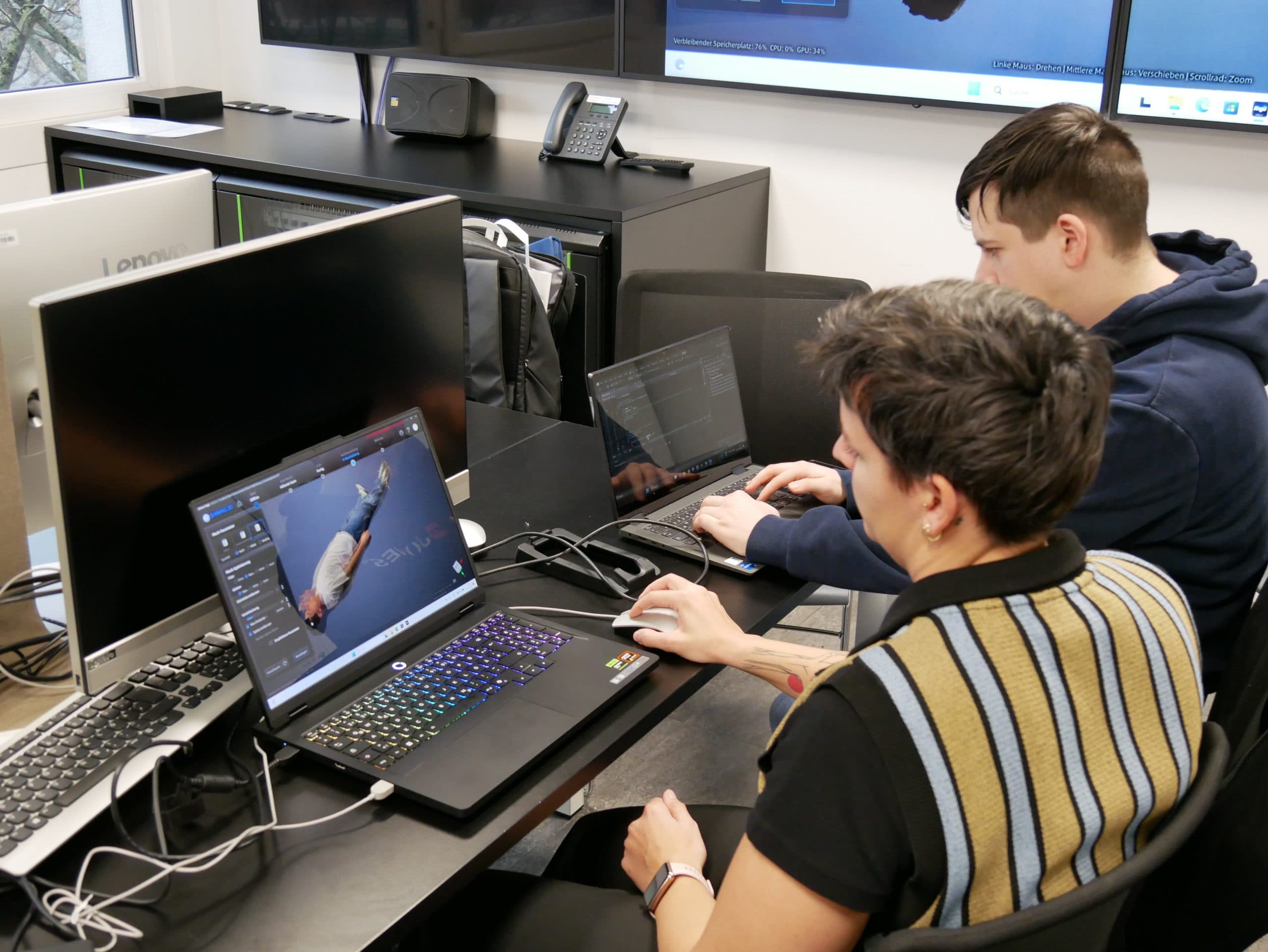 Zwei Personen sitzen an Schreibtischen mit Laptops und bearbeiten konzentriert ein 3D-Modell auf einem Bildschirm. In einem modernen Büro oder Klassenzimmer sind mehrere Monitore und Computergeräte zu sehen.