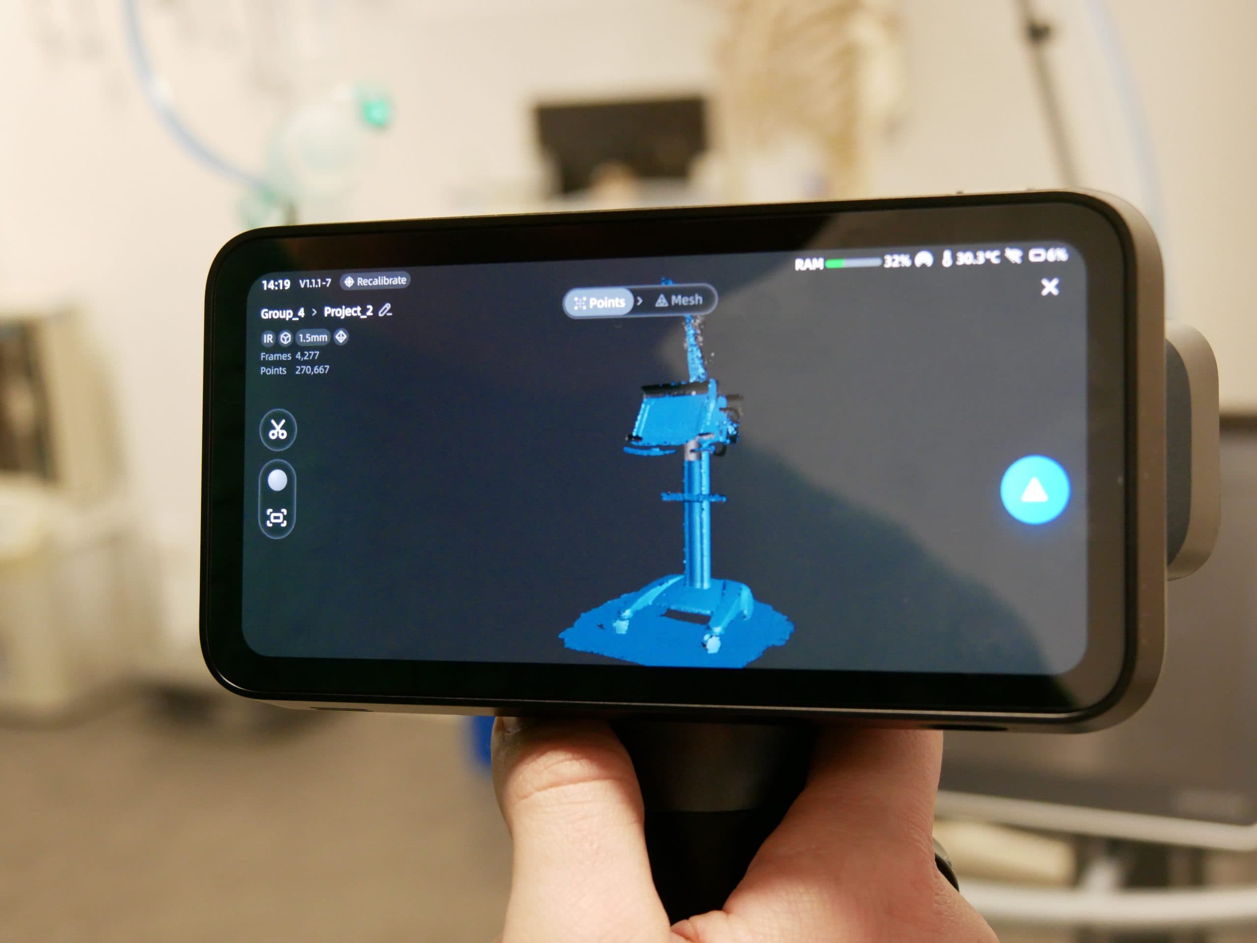 Eine Hand hält ein Smartphone, das einen 3D-Scan einer medizinischen Ausrüstung anzeigt, wobei auf dem Bildschirm blaue Punktwolkendaten zu sehen sind. Der Hintergrund zeigt eine unscharfe Labor- oder Klinikumgebung.