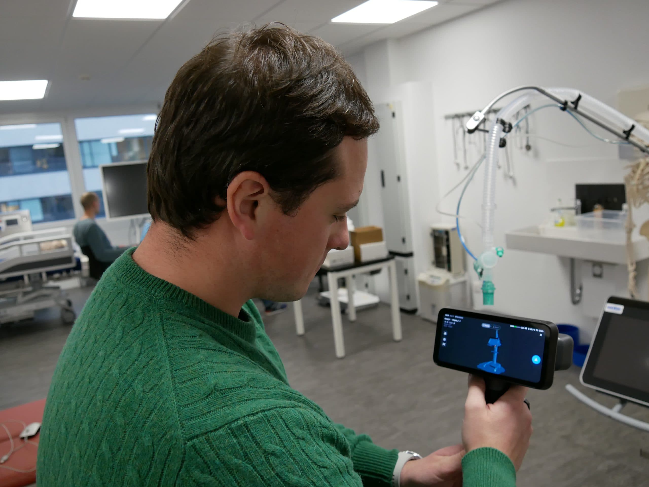 Ein Mann in einem grünen Pullover hält ein Smartphone in der Hand, auf dem ein blaues technisches Diagramm in einem modernen medizinischen Labor zu sehen ist, mit Geräten und einer weiteren Person im Hintergrund.