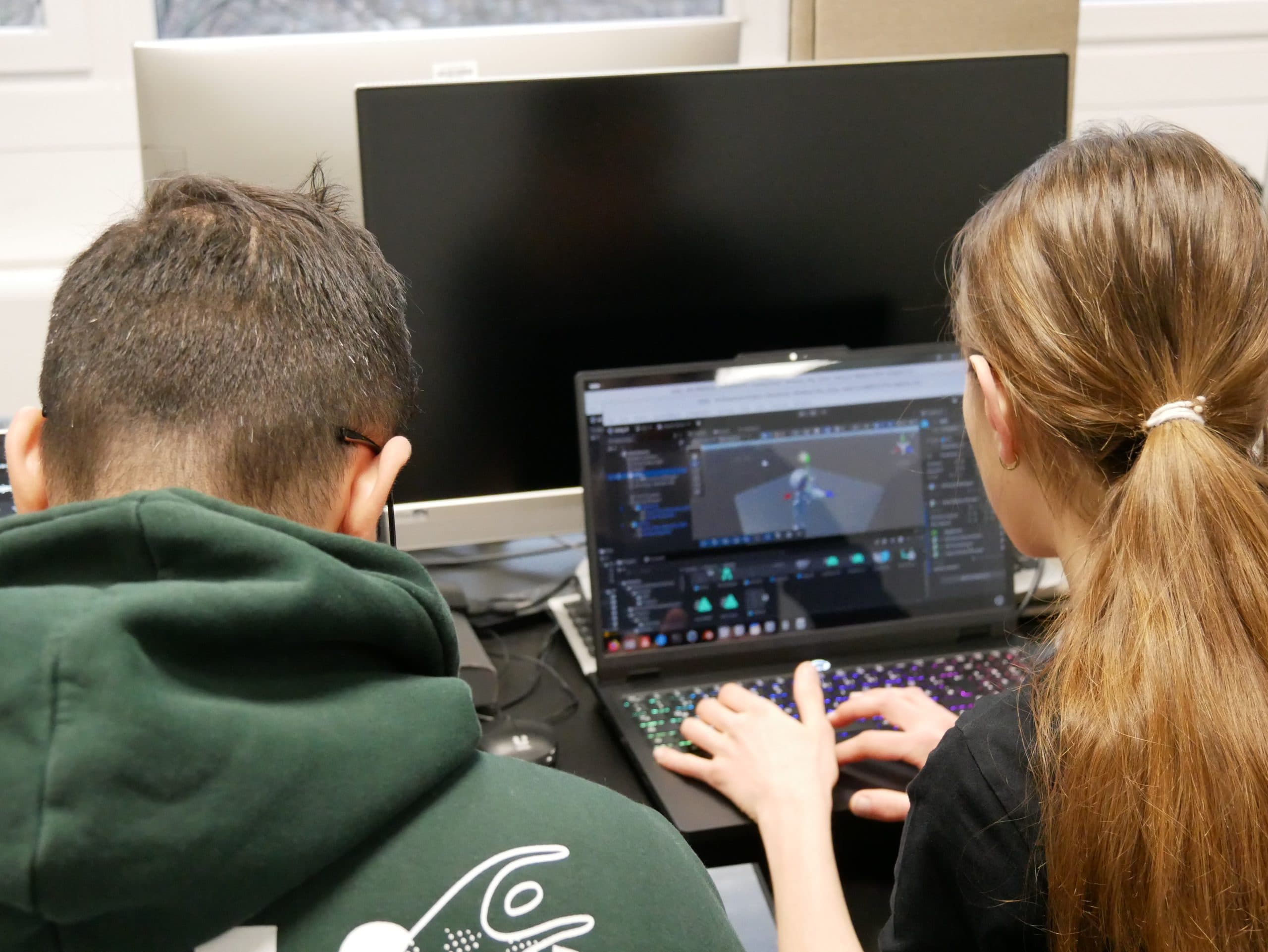 Zwei Personen sitzen nebeneinander an einem Schreibtisch und konzentrieren sich auf einen Laptop mit einer 3D-Modellierungssoftware. Vor ihnen befindet sich ein großer, ausgeschalteter Monitor. Die Szene scheint eine gemeinschaftliche Arbeits- oder Lernumgebung zu sein.