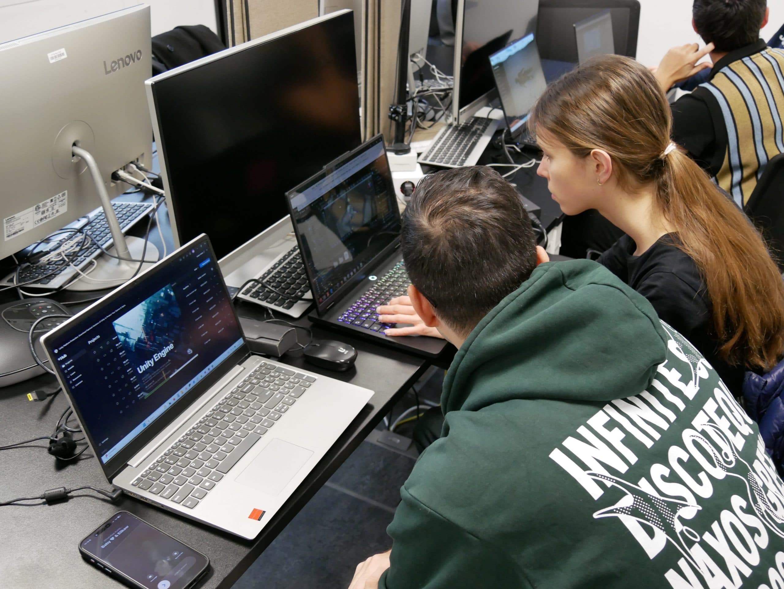 Zwei Personen sitzen an einem Schreibtisch mit Laptops und konzentrieren sich auf ihre Bildschirme. Einer trägt einen grünen Kapuzenpullover mit weißer Schrift, der andere hat langes braunes Haar. Um sie herum sind mehrere Computer, Monitore und Kabel in einem geschäftigen Arbeitsbereich zu sehen.