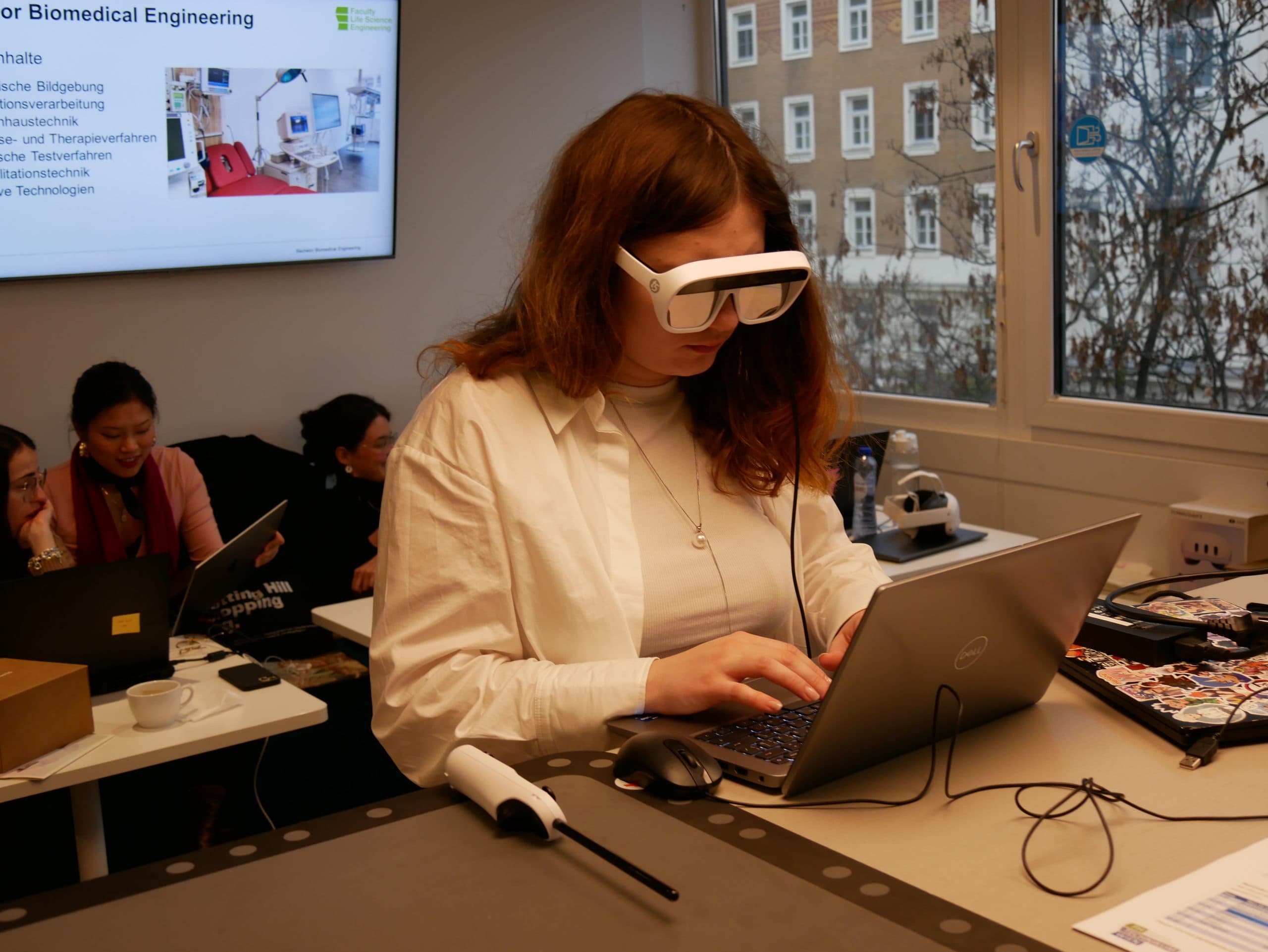 Eine Frau mit einer Augmented-Reality-Brille arbeitet in einem Klassenzimmer an einem Laptop. Im Hintergrund sind andere Schüler und eine Präsentationsfolie über biomedizinische Technik zu sehen.