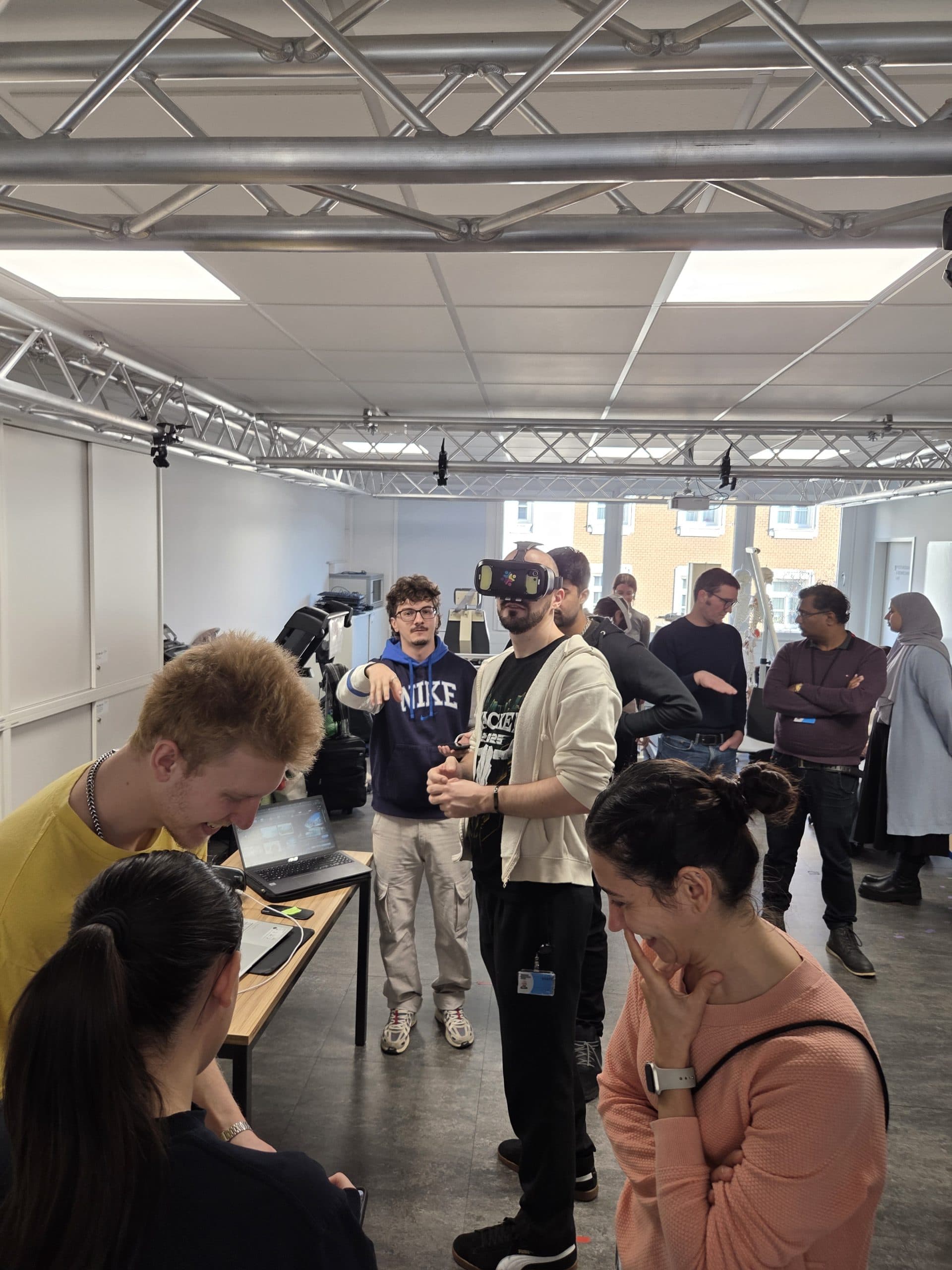 Eine Gruppe von Menschen ist in einem Raum mit technischen Geräten versammelt. Eine Person trägt ein VR-Headset, während andere um einen Tisch herum beobachten oder interagieren. Der Raum hat große Fenster und Deckenleuchten aus Metall.