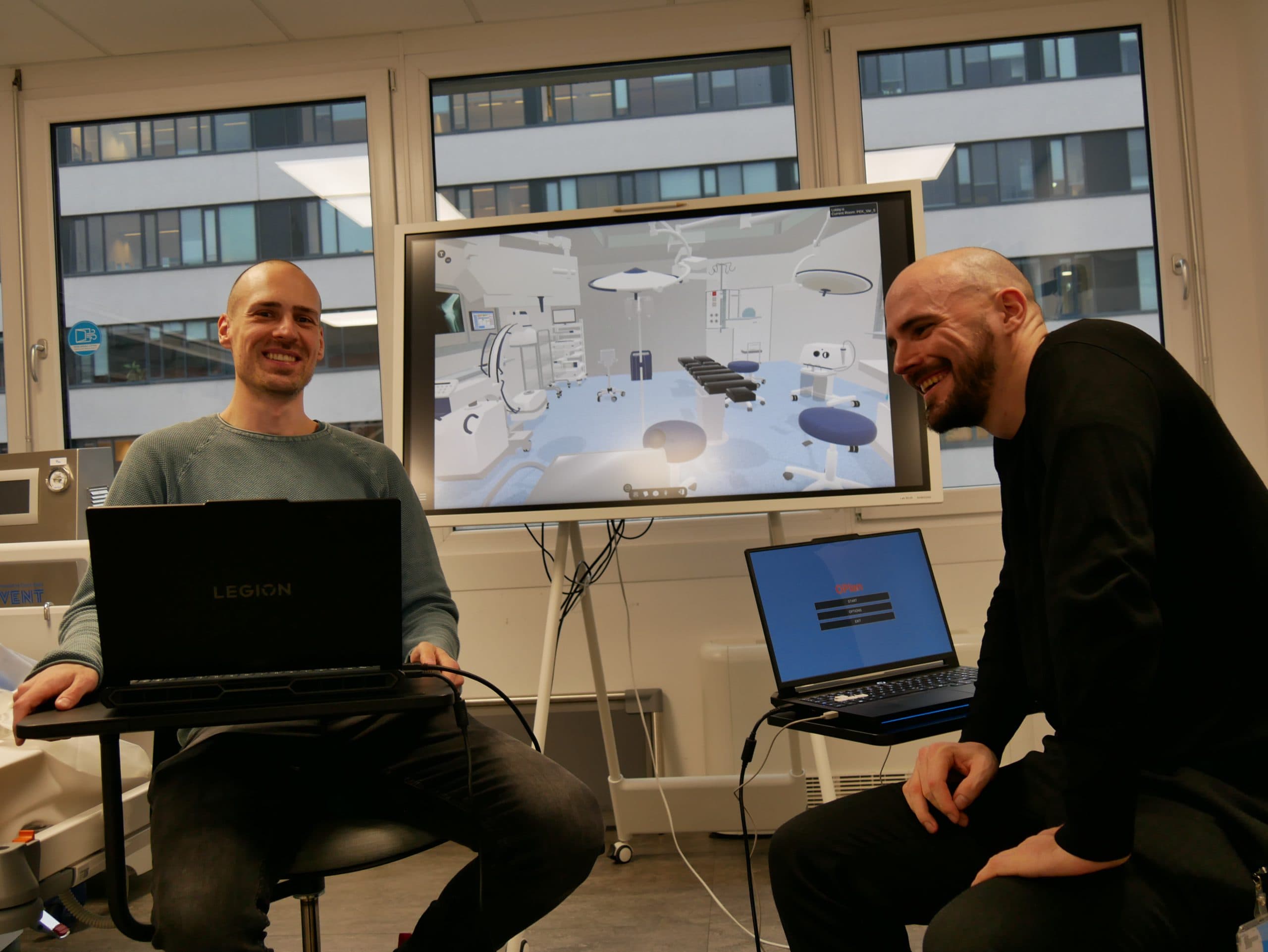 Zwei lächelnde Männer sitzen mit Laptops in einem modernen Büro. Hinter ihnen zeigt ein großer Bildschirm ein digitales 3D-Modell eines Operationssaals. Große Fenster zeigen ein Bürogebäude im Freien.