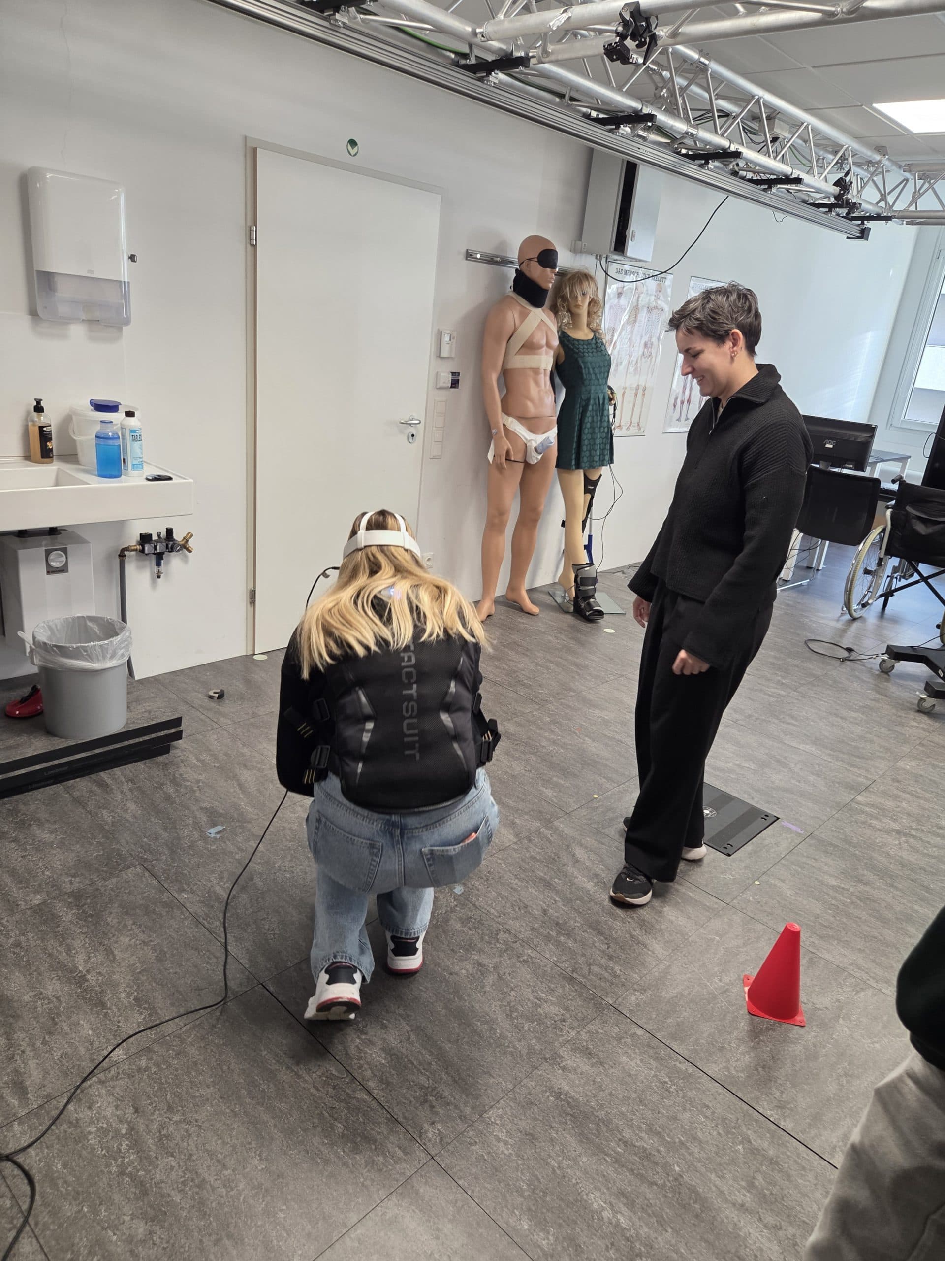 Eine Person hockt auf dem Boden und hantiert mit Geräten, während eine andere in der Nähe steht und lächelt. Im Hintergrund befinden sich zwei lebensgroße Schaufensterpuppen, die als Patienten gestaltet sind, ein Waschbecken und verschiedene medizinische Geräte.