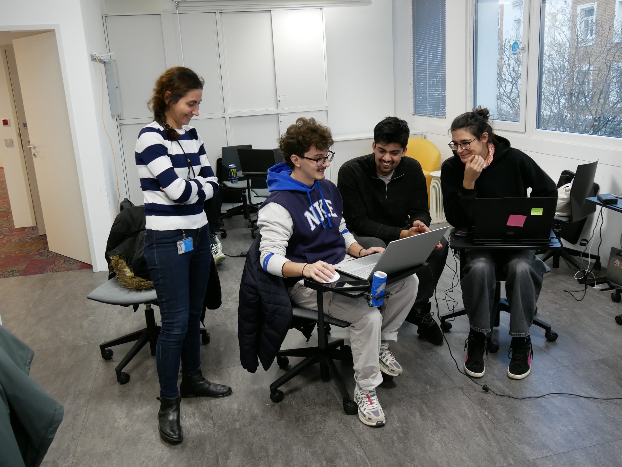 Vier Personen sind in einem Büro versammelt. Zwei sitzen an Schreibtischen mit Laptops, während die anderen beiden stehen und zusehen. Alle lächeln und scheinen zusammenzuarbeiten oder eine Diskussion zu genießen.
